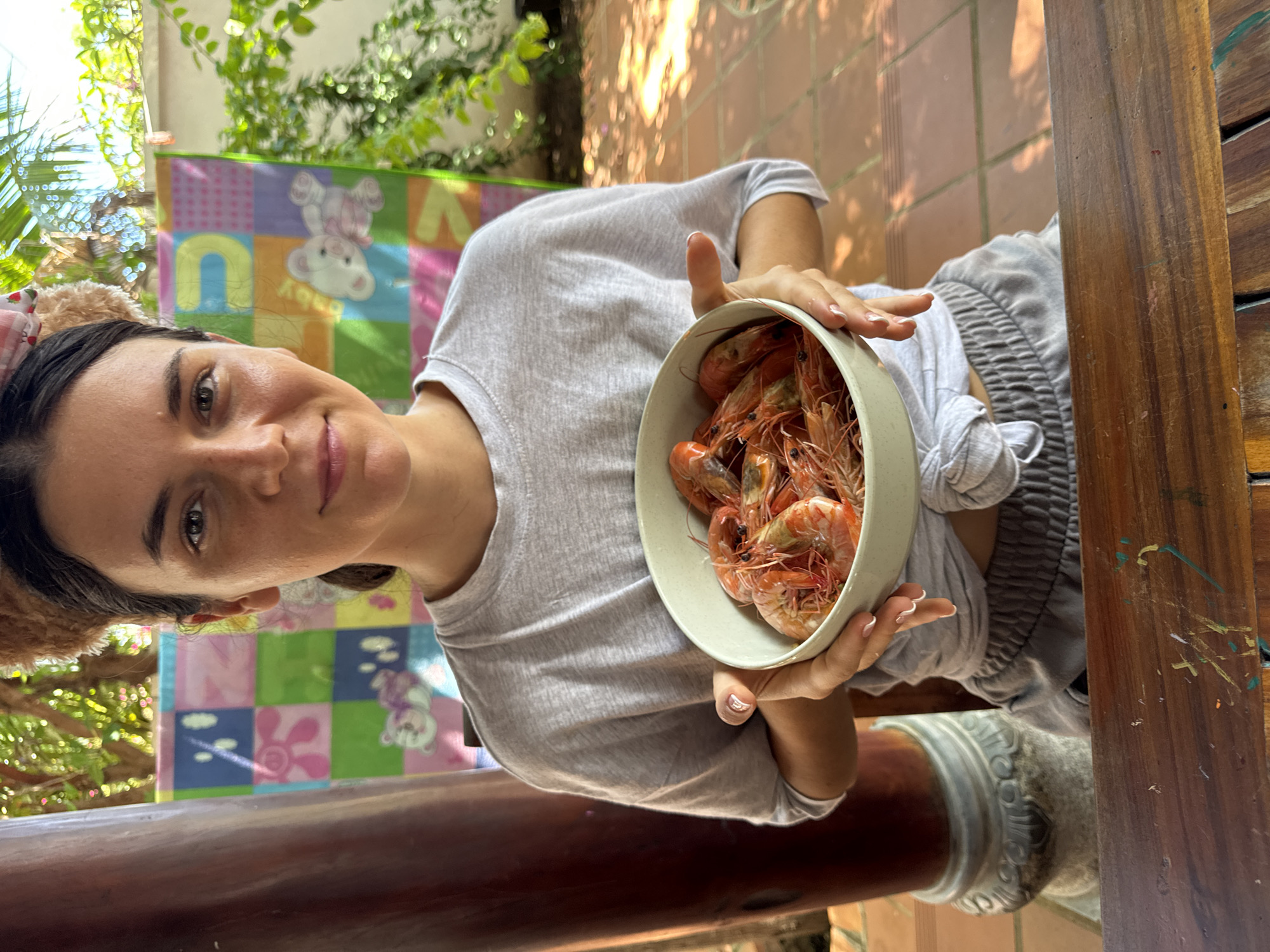 A woman holds a bowl of cooked shrimp in an outdoor setting in Mũi Né, Vietnam. She faces the camera with a slight smile, positioned to the left of the bowl. Her attire consists of a gray t-shirt and gray pants, possibly pajama pants. The bowl, off-white, is filled with reddish-brown cooked shrimp.
The background includes a child's playmat visible behind her head. Sunlight filters through trees overhead, casting shadows on the scene. The setting is on an outdoor patio with tiled flooring. A wooden structure is visible to the right of the woman.