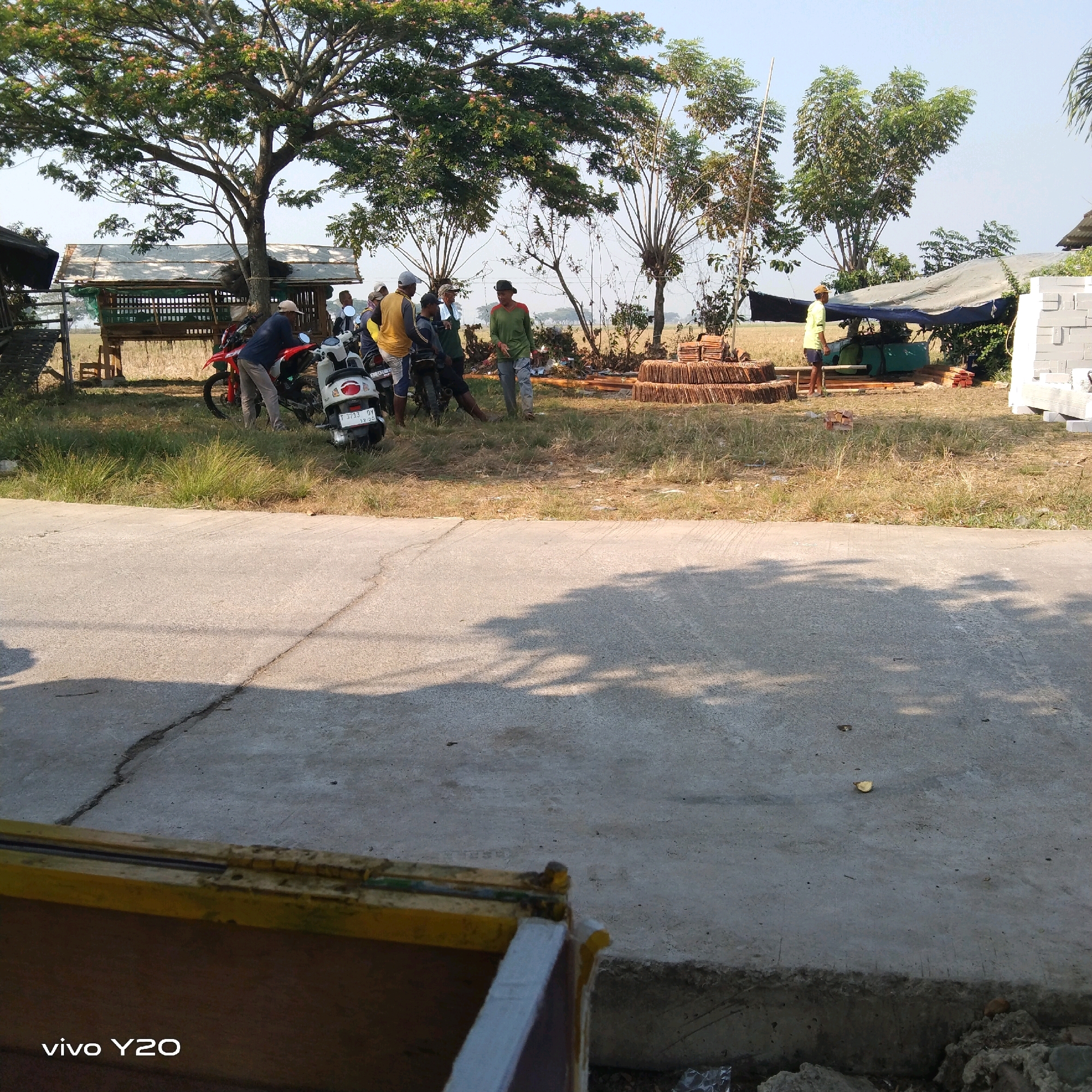 In an outdoor rural setting, a group of men are gathered around motorcycles and what appears to be a construction or material processing site. The scene includes several men, some standing and others near motorcycles. One man is wearing a yellow shirt and blue shorts, another a green long-sleeved shirt and grey pants, and others are in casual clothing. Two motorcycles, one red and one white, are parked on a grassy area adjacent to a paved road.
The background features a modest wooden structure with a white tarp roof, several trees, and stacks of what appear to be clay tiles or bricks. To the right, a large tarp is draped over equipment, and more building materials, including white blocks, are visible. The environment is dry with sparse vegetation and a clear sky. The foreground shows a cracked paved surface with shadows cast by overhead trees. The "vivo Y20" watermark is present in the bottom left corner, indicating the device used for capture.