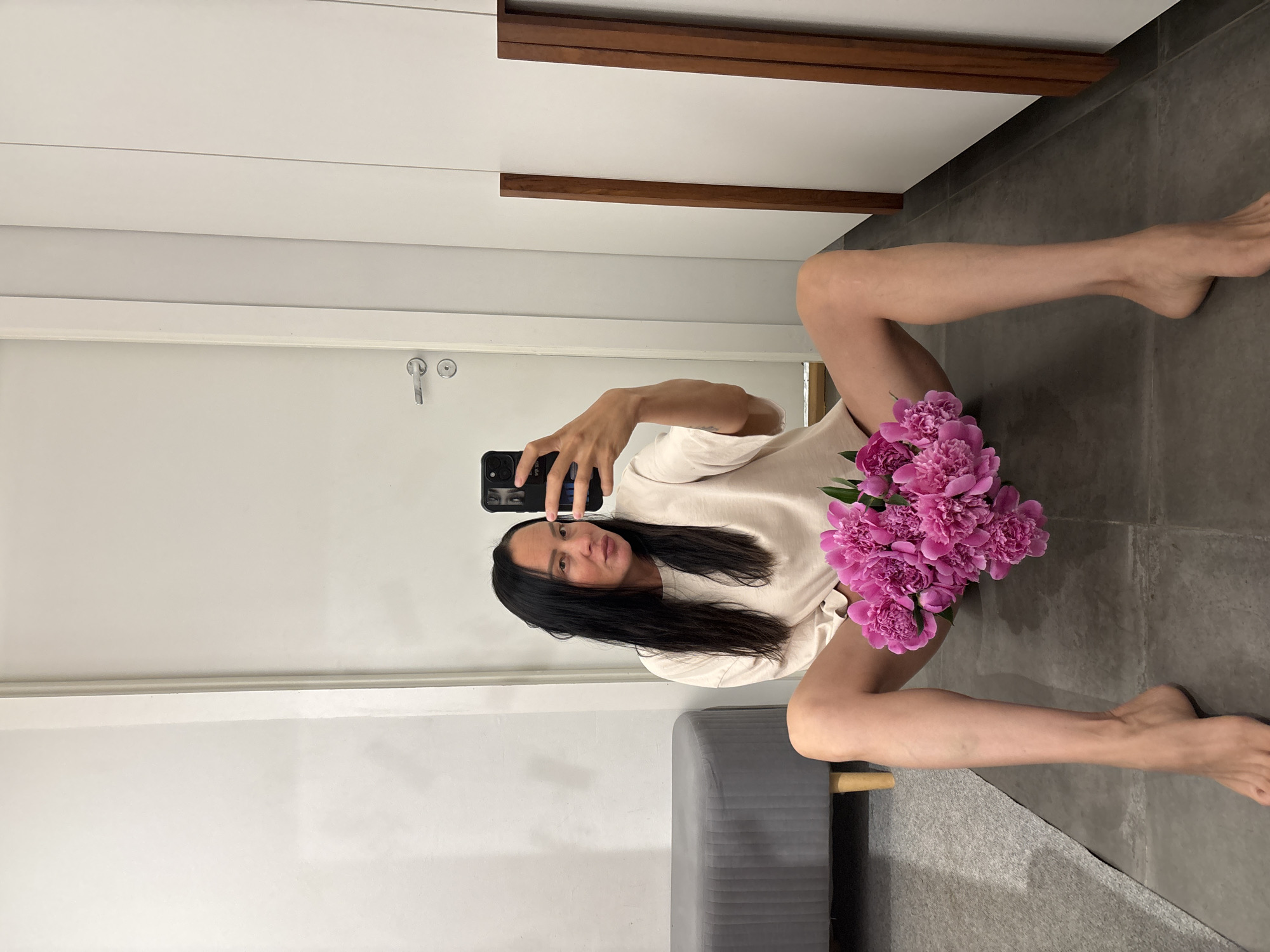 A woman is taking a selfie in a room. She is sitting on the floor with her legs spread and a bouquet of pink peonies in her lap. She is wearing a light-colored t-shirt. Her phone is in her right hand and she is looking at the camera. The room has a grey floor and white walls. There is a grey ottoman visible and a white closet with wooden accents. The photo was taken in the city of Chaikovsky, Russia.