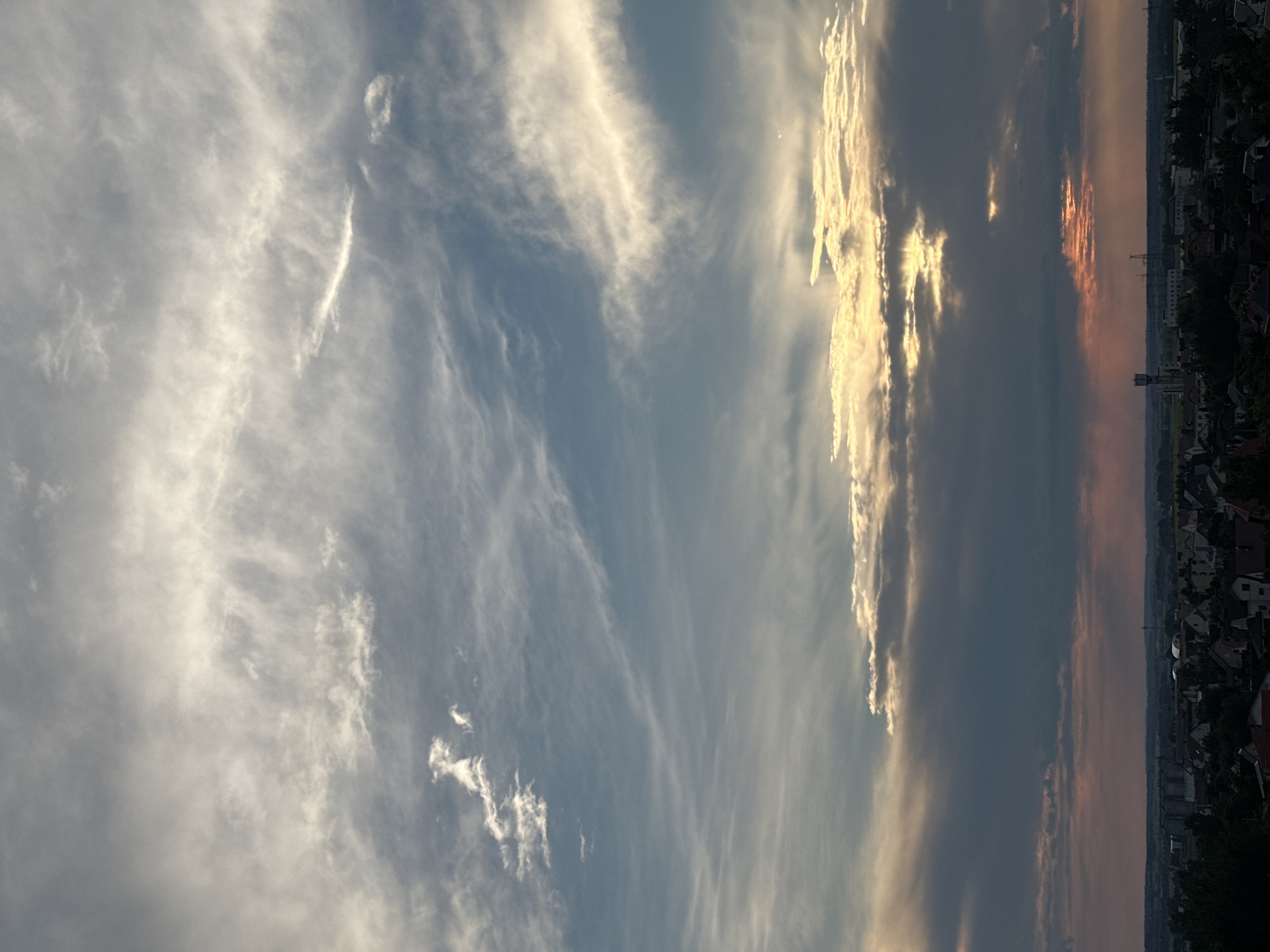 The image depicts a broad, panoramic view of the sky above the Шчомысліцкі сельскі Савет in Belarus. The sky is filled with a dramatic display of clouds, with bands of cirrus clouds streaking across a pale blue background in the upper portion of the frame. Lower down, thicker, darker clouds are present, some illuminated by the sun breaking through in golden shafts. The sun itself is partially obscured by these clouds, creating a sunset or sunrise effect with warm tones of yellow and pink visible near the horizon. Below the sky, the distant landscape is visible, featuring a low-lying urban or suburban area with numerous rooftops, trees, and some industrial structures, including a tall, slender tower. The overall atmosphere is one of a dynamic and visually striking sky over a settled area.