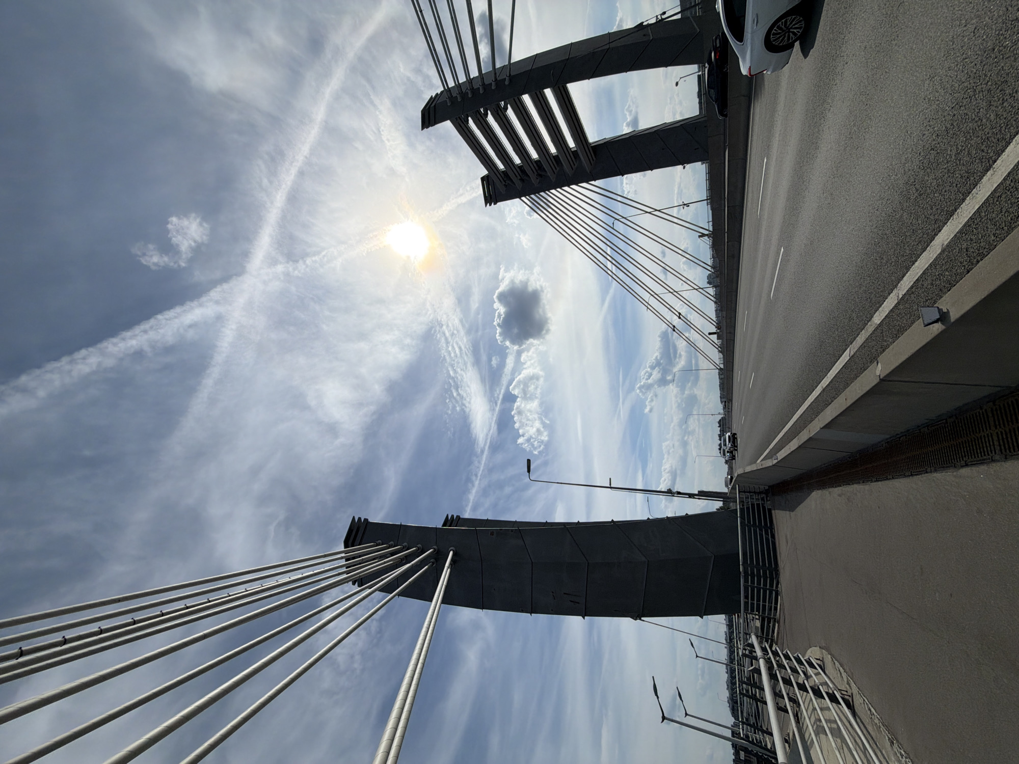 This is a low-angle, wide-angle photograph depicting the central section of the Western High-Speed Diameter (WHSD) bridge over the Korabelny Fairway in Saint Petersburg, Russia, observed on a clear day with scattered clouds.
The scene is dominated by the structural elements of the cable-stayed bridge. Two dark grey, geometrically faceted concrete pylons stand prominently against the sky. The pylon on the immediate left foreground is closer and more fully visible, exhibiting a tapering design. The second pylon is situated further along the bridge on the right side of the frame, partially obscured by its distance and the overall perspective. Numerous white-grey tension cables extend from the upper sections of both pylons downwards, connecting to the unseen bridge deck level, creating strong diagonal lines across the frame.
A multi-lane asphalt highway occupies the central and right portions of the image, showing clear white lane markings. A concrete barrier delineates the highway from a narrower concrete pedestrian or maintenance path on the left foreground. This path is bordered by a metal guardrail consisting of multiple horizontal bars.
Vehicular traffic is present on the highway. A white sedan is partially visible in the far right lane in the immediate foreground, with its rear portion and right side wheel showing. Further down the road, a white SUV or van is visible, and a dark sedan is also observable under the distant right pylon. No human figures are discernible on the bridge.
The sky is bright blue with scattered white stratocumulus clouds. Multiple high-altitude contrails are visible, with two prominent contrails intersecting to form an 'X' shape in the upper-mid section of the sky. The sun, partially diffused by thin clouds, is a bright, overexposed light source in the upper-right quadrant. A distinct, isolated cumulus cloud is situated in the upper-left sky section, near the left pylon. Modern black streetlights with angled luminaires are positioned along the bridge structure.