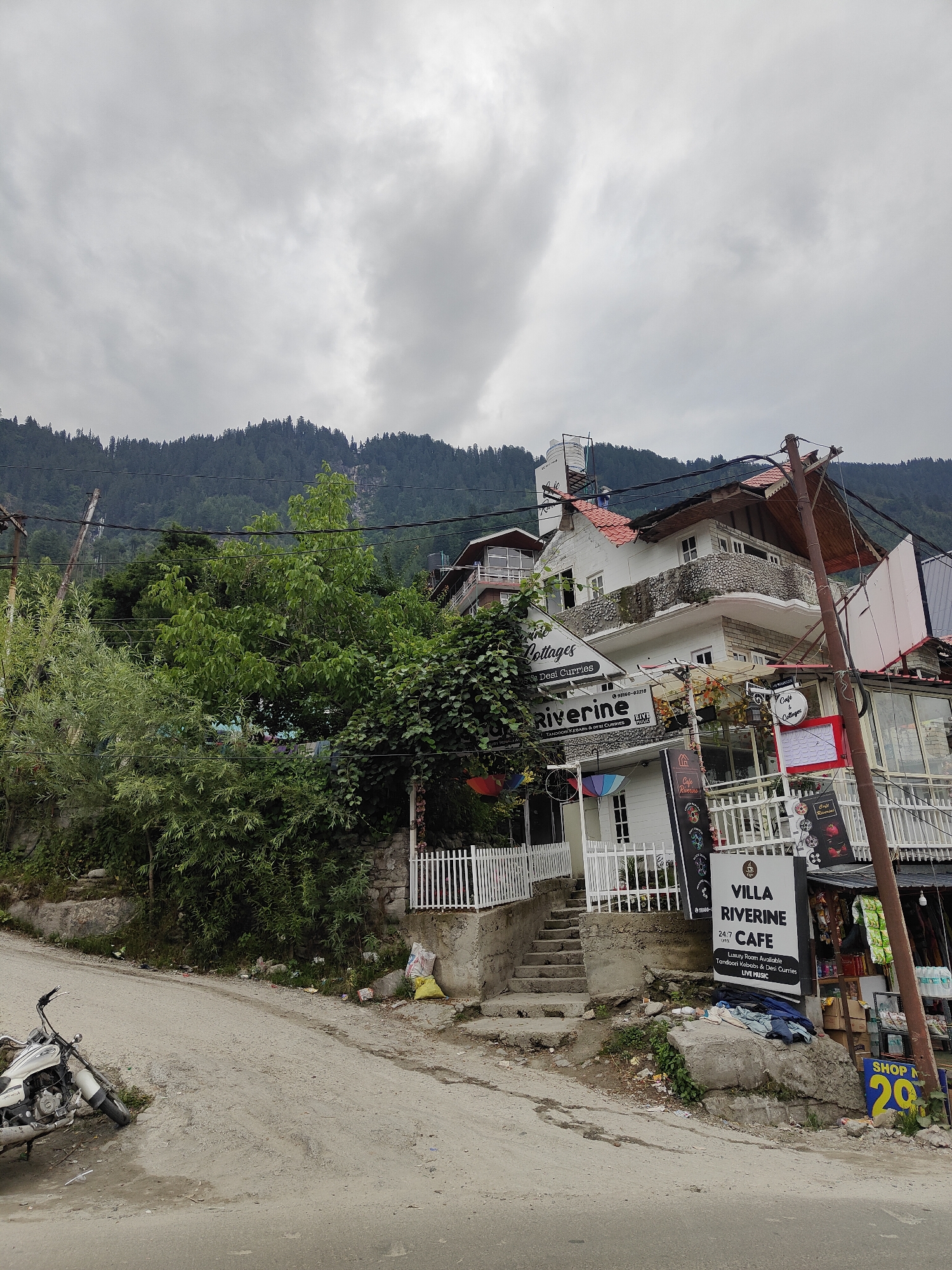 A white motorcycle is parked on the left side of a dusty, uphill road in Vashisht, India. A cluster of multi-story buildings with white exteriors and red-tiled roofs are situated on the right side of the road, ascending the hill. Several signs are visible on the buildings, including "Cottages," "Riverine," and "VILLA RIVERINE CAFE." A white picket fence borders the property in front of the buildings. Lush green trees and foliage surround the buildings and grow on the hillside. In the background, a dense forest covers a steep mountain under a cloudy sky. The overall scene depicts a bustling area with a mix of residential and commercial establishments nestled within a mountainous landscape.