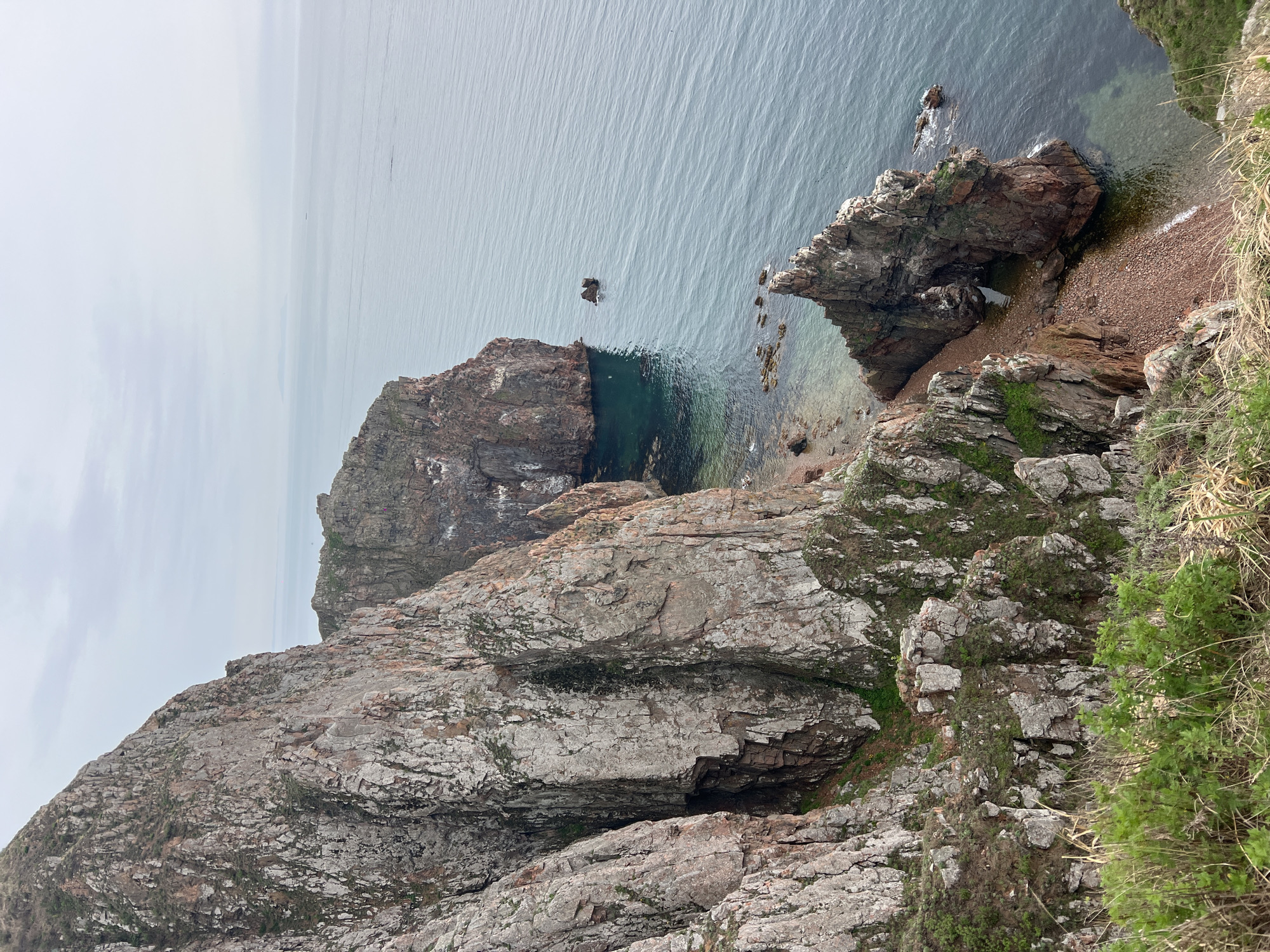 This image presents a top-down view of a rocky coastline, likely located in a coastal region. The scene is dominated by various shades of grey and brown rocks, some of which are partially covered with green vegetation. The water is calm and a muted grey-blue, with some areas appearing more transparent near the shoreline, revealing rocky formations below. There is a small beach with reddish-brown pebbles visible near the rocks. A rock formation is seen extending into the sea, featuring a small archway. The sky is overcast. The location is Russkiy Island, Russia.