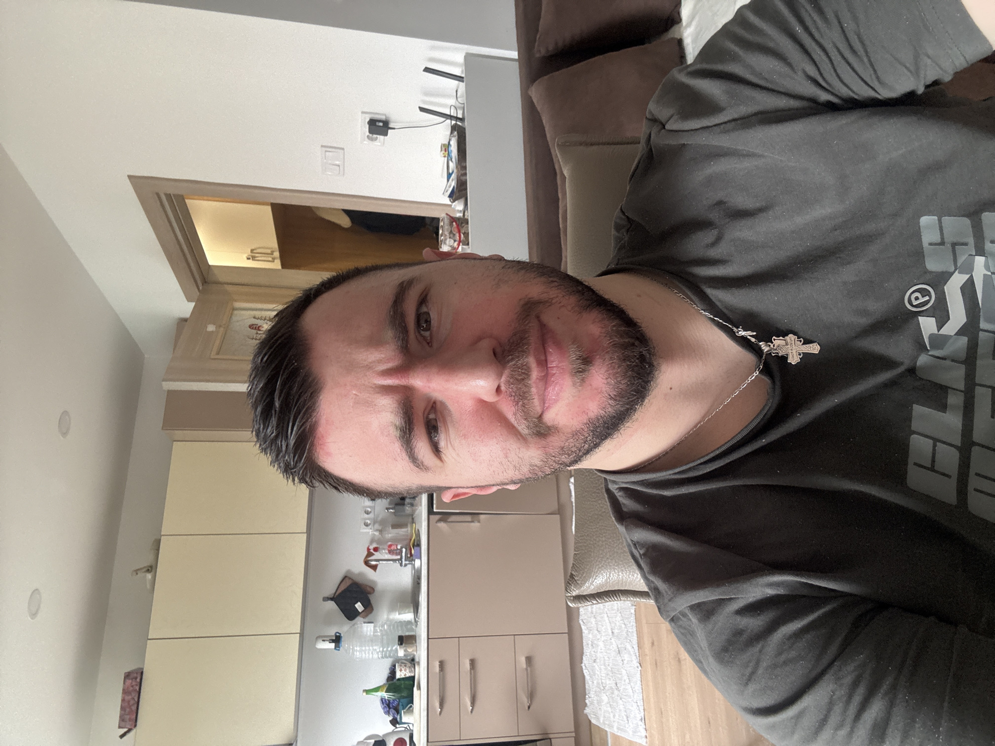 A man is depicted in a selfie-style photograph, likely taken indoors. He has dark hair, a beard, and is wearing a dark-colored t-shirt with a silver necklace featuring a cross pendant. The background shows elements of a modern interior. In the background, beige kitchen cabinets are visible. There is also an open doorway leading to another room with brighter lighting. A portion of a sofa with brown pillows is seen to the right. The picture does not provide enough information to determine that the scene is located in the city of Sofia, Bulgaria.
