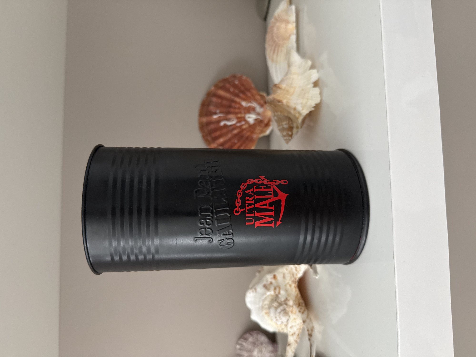 A black, cylindrical container with horizontal ridges is positioned on a white shelf. The container has text and an emblem printed in red. The text reads "Jean Paul Gaultier" at the top and "ULTRA MALE" with an anchor symbol below it. The background is a plain, light-colored wall. Several seashells are arranged around the container on the shelf. The image does not contain any information that would allow for its localization in Burgas, Bulgaria, or any other specific city.