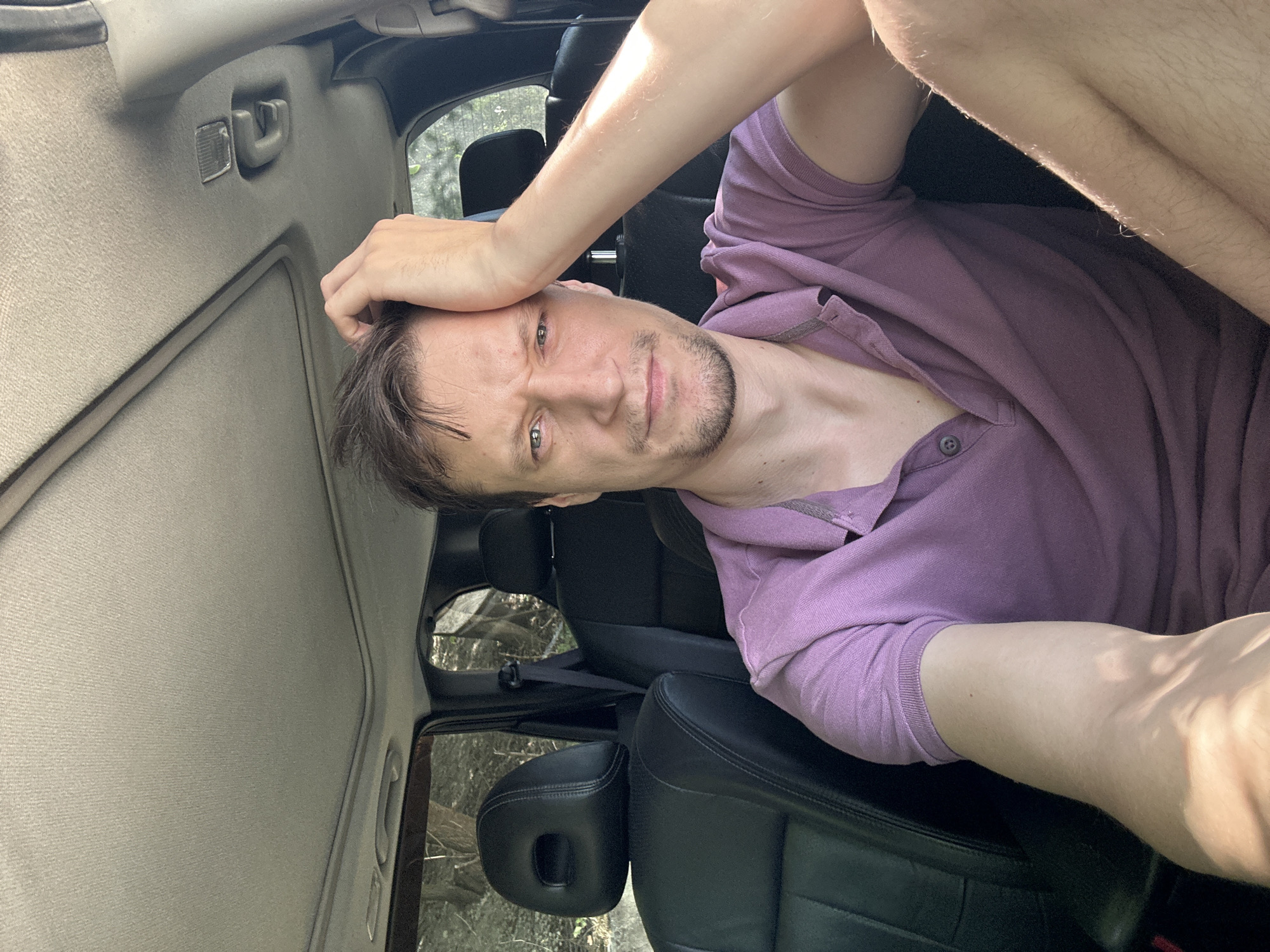 A man with brown hair and a short beard, wearing a purple polo shirt, is sitting in the driver's seat of a car. He is holding his left hand to his forehead and looking at the camera with a pensive expression. The interior of the car, including the black leather seats and the sunroof, is visible. The background suggests an outdoor environment, possibly a wooded area. The image appears to be a selfie taken in daylight.