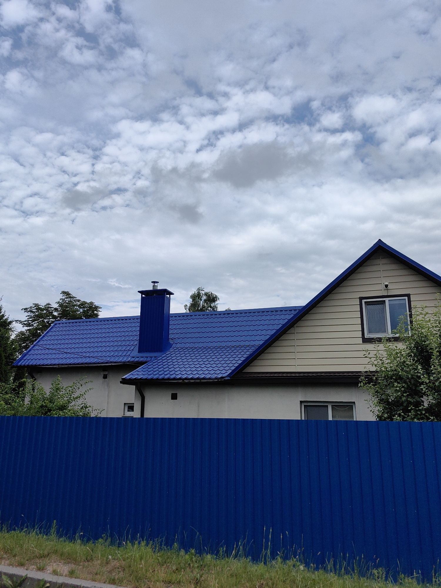A residential house with a bright blue metal roof and a matching blue chimney is pictured on a partly cloudy day. The house has beige siding and white walls, with a few windows visible. A tall, solid blue corrugated metal fence runs across the foreground, obscuring the lower portion of the house. Lush green trees and bushes surround the property, adding to the natural setting. The sky is filled with a mixture of white and gray clouds, suggesting a temperate climate. The scene appears to be in a suburban or rural area, possibly in Fanipal, Belarus, given the overall aesthetic and foliage. No people or specific activities are depicted, but the well-maintained appearance of the house and fence suggests a lived-in home. The time of day is likely daytime, given the light illuminating the scene.
