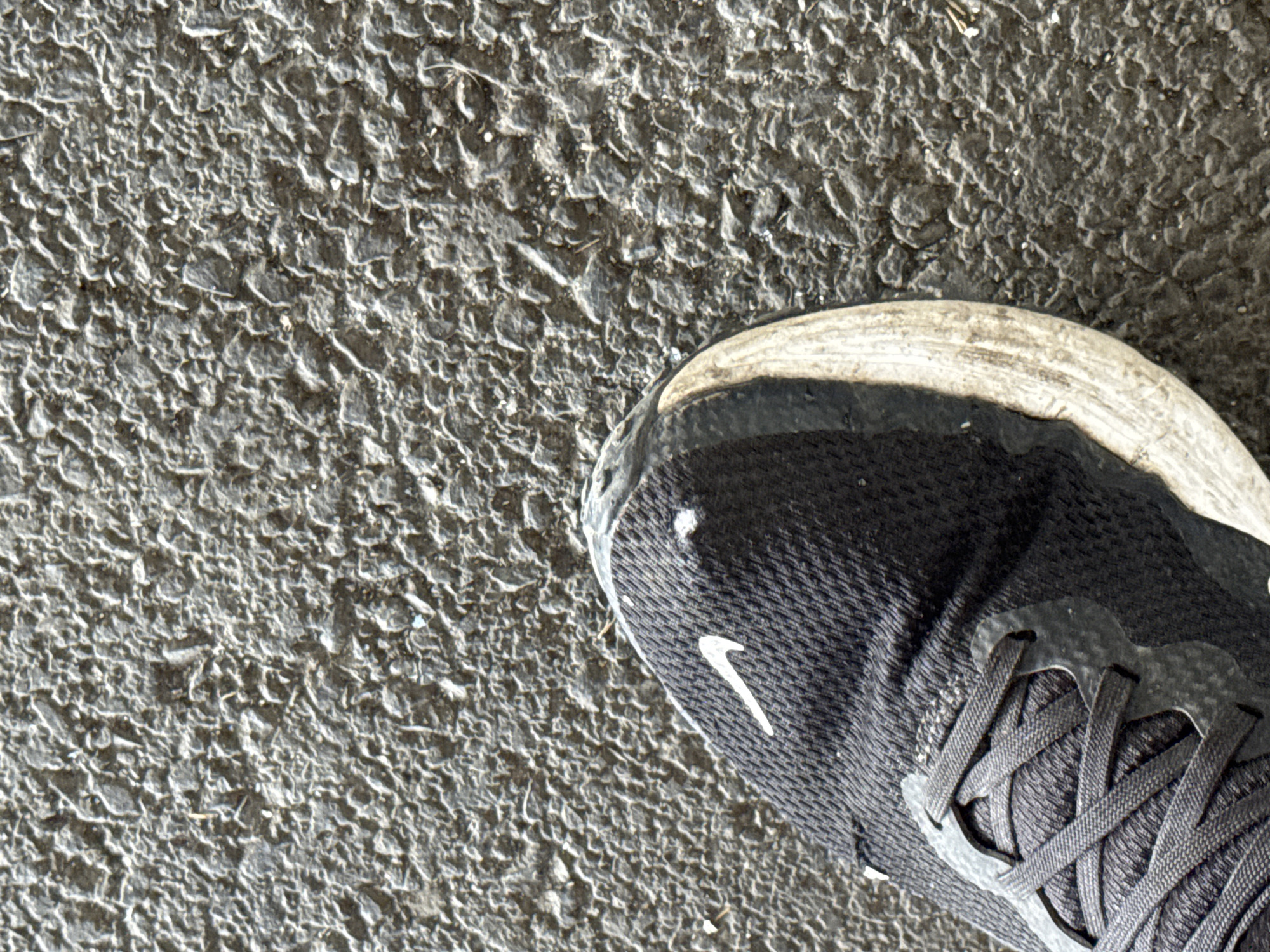 A close-up, high-angle shot shows the toe of a black athletic shoe with a white Nike swoosh logo against a rough, dark gray asphalt surface. The shoe's white sole is partially visible and appears dirty. Small white speckles are present on the shoe and the asphalt. The scene is likely outdoors in an urban environment, possibly a street or sidewalk in Burgas, Bulgaria, given the characteristic texture of the asphalt. No people are visible, and the focus is solely on the shoe and the ground.