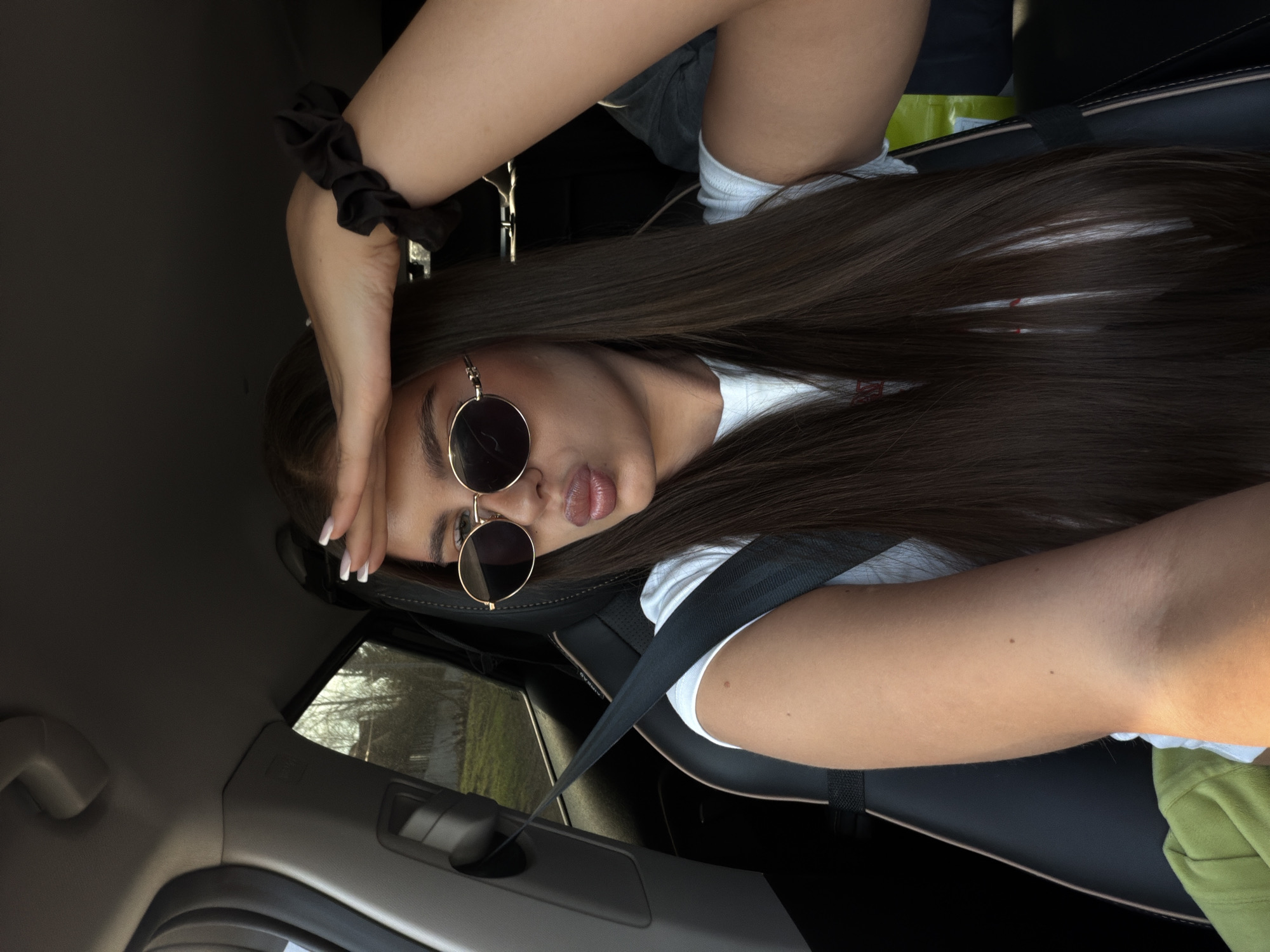 A young woman sits in the driver's seat of a car, wearing sunglasses and a seatbelt. She is looking at the camera with a pout. Her left hand rests on her forehead, a black scrunchie on her wrist. The car's interior is visible, as is a glimpse of a building exterior through the window. The location is potentially Krasnoyarsk, Russia, based on contextual information.