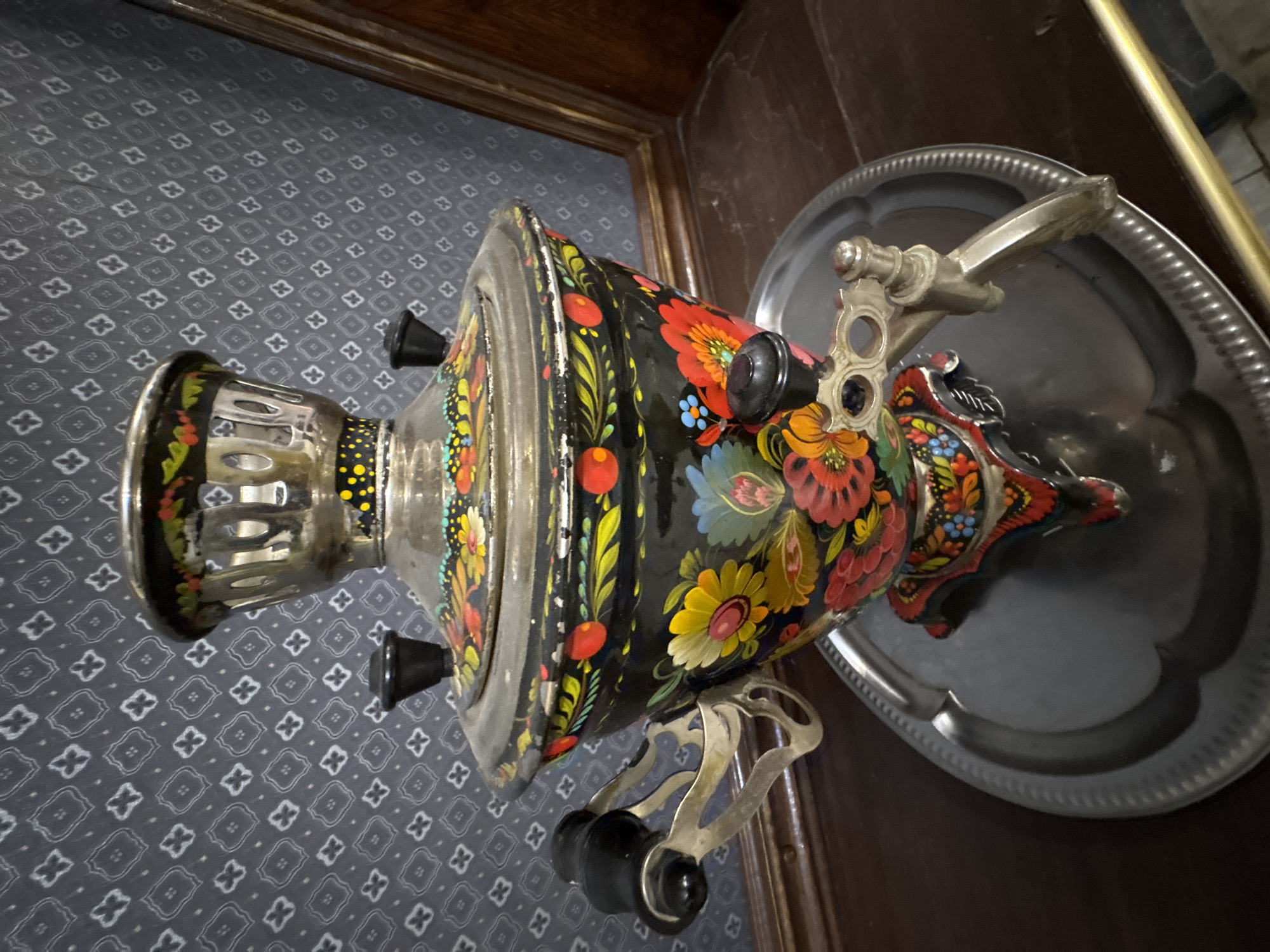 A close-up, overhead shot focuses on a decorative, painted samovar, likely an antique, placed on a silver tray. The samovar is predominantly black with vibrant, folk-art style floral patterns in red, yellow, blue, and green. It has a prominent spout on the right side and two black handles with metal accents on the sides. The background features a dark, patterned fabric, possibly a tablecloth or wallpaper, with a repeating geometric design. A wooden frame is visible at the top left of the image. The lighting suggests an indoor setting, with no clear indication of the time of day or weather. The image appears to be taken in Zavod Mikhailovskii, Russia, given the location context. No text is visible in the media file.