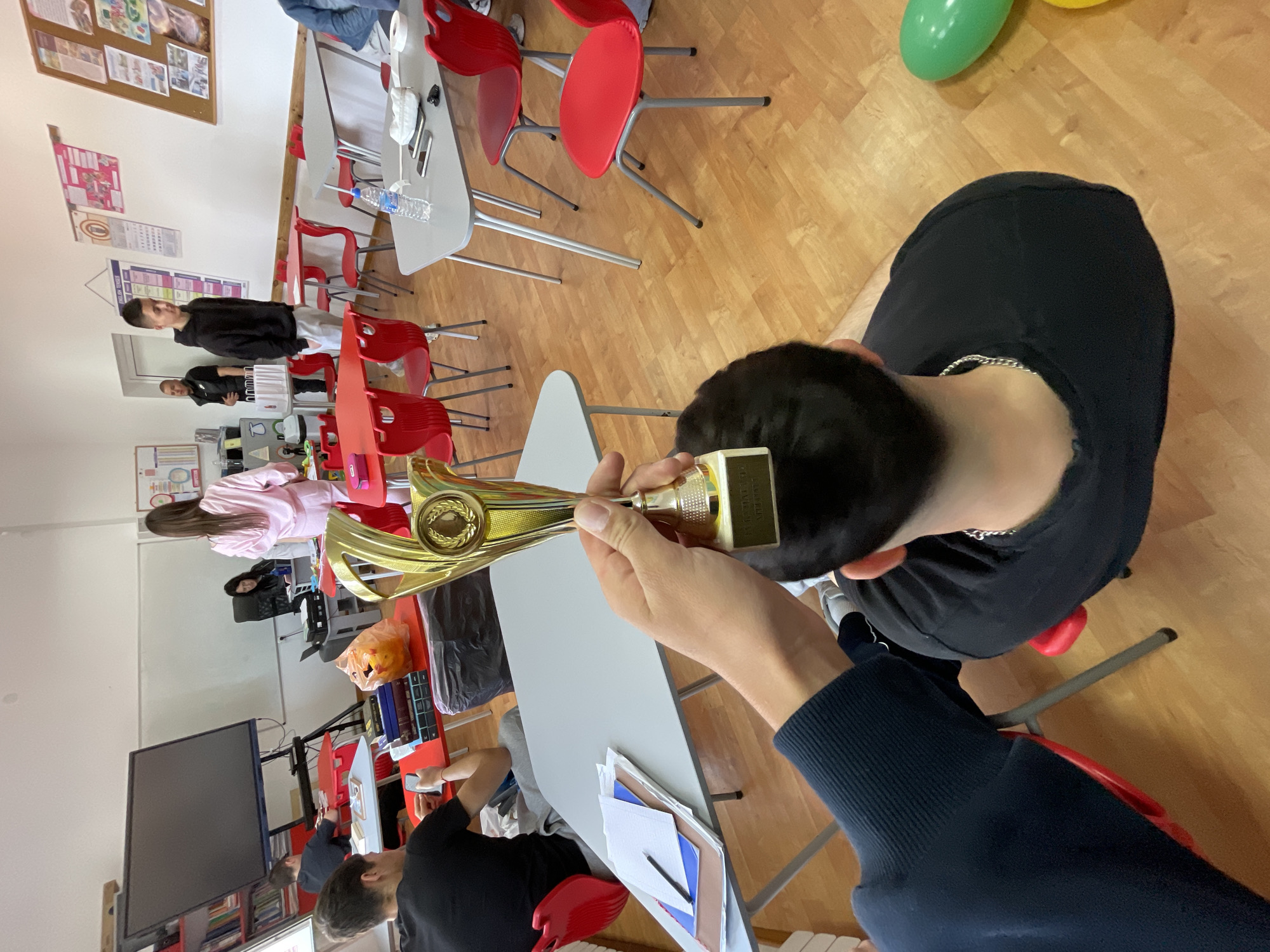 A person is holding a gold trophy in an interior space. The person is wearing a black shirt and a dark blue long-sleeved shirt. They are sitting on a red chair at a gray table covered in papers and writing implements. In the background, people are seated at tables, some of which are red. The floor is wooden. There is a large television screen on a wall to the left and a bulletin board on a wall in the background. There are red chairs and gray tables. The location is potentially Zlatograd, Bulgaria.