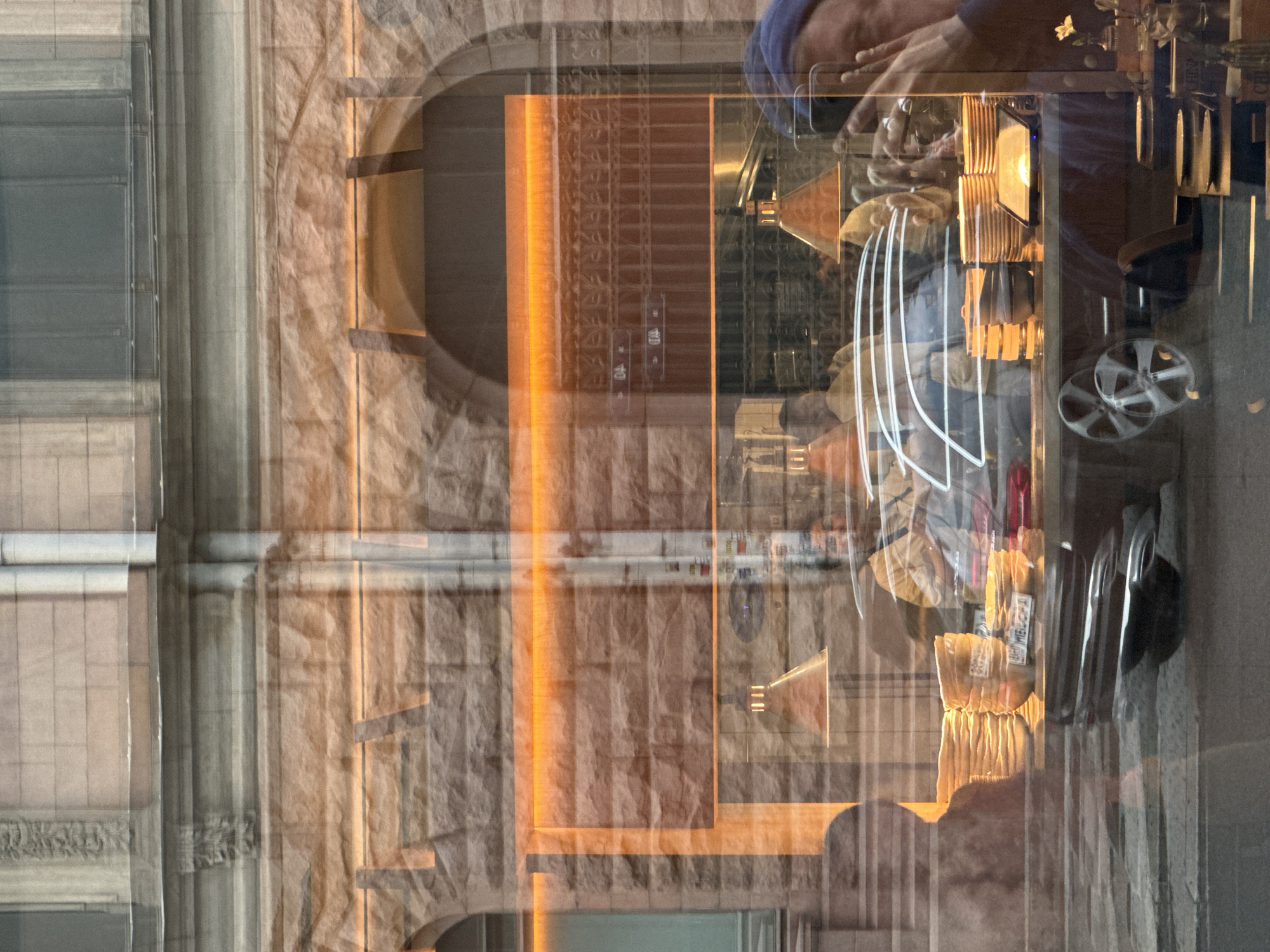 This is a reflective shot of a street scene in Saint Petersburg, Russia, viewed through a window. The window itself is a prominent subject, displaying reflections of the building opposite, which features stone masonry, arched doorways, and a horizontal orange light fixture.
Inside the window, a cafe or restaurant is visible. Several stacks of plates are arranged on a counter or shelf, some with labels. There are multiple people inside, appearing as silhouettes and blurred figures due to the reflections and glass. One individual, towards the right, is holding a smartphone and taking a picture of themselves, wearing a blue hat. Another person, to the left, is mostly a silhouette. The interior also shows some lighting fixtures, one prominently an orange conical lamp. A black car is parked or driving by, its reflection visible on the street level. The overall impression is one of a busy urban environment with layers of reflection creating a complex visual tapestry.