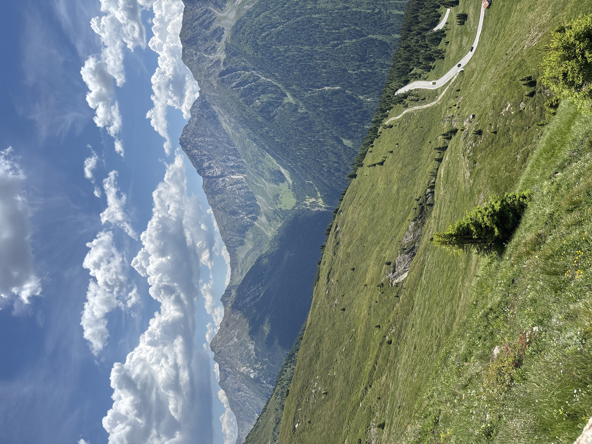 This is a landscape photograph taken in the mountains of St. Leonhard in Passeier - San Leonardo in Passiria, Italy. The image depicts a winding road with several cars and a bus, snaking its way up a lush green mountain slope. The road is bordered by dense coniferous trees on one side and a grassy slope with scattered small trees and rocks on the other. In the background, majestic, rugged mountain peaks rise against a bright blue sky dotted with fluffy white clouds. The overall scene conveys a sense of natural beauty and the vastness of the mountainous terrain.