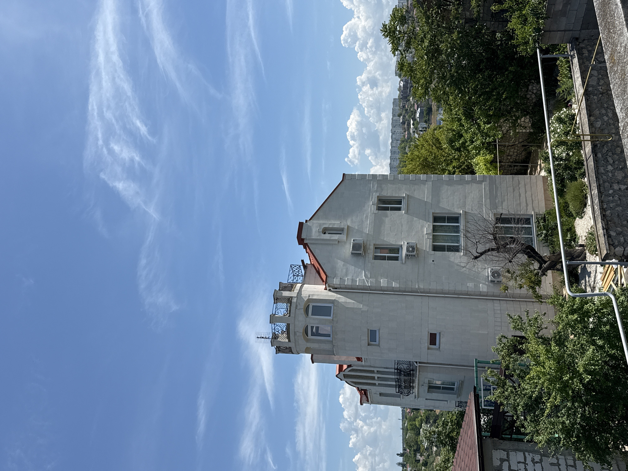This detailed image captures an outdoor scene under a clear blue sky with various cloud formations, depicting a residential area in the city of Unknown, Unknown. The central focus is a multi-story, light-colored building with a distinctive architectural style, featuring a prominent round tower on its left side topped with a decorative dark metal balustrade and an antenna. The building has a red-tiled roof, multiple windows (some arched), and dark ornate metal balconies. Several exterior air conditioning units are affixed to its walls.
In the mid-ground, to the right of the main building, a cluster of larger, multi-story residential buildings is visible on a distant hillside, suggesting a broader urban landscape. The foreground and immediate surroundings are characterized by lush green trees and dense foliage. A partially constructed wall made of concrete blocks is visible in the lower left, and a metal railing extends across the bottom of the frame, indicating an elevated vantage point. Below this railing, a garden area with small shrubs, white flowers, and light-colored gravel is visible, alongside a bare, twisted tree. No people are present in the scene.