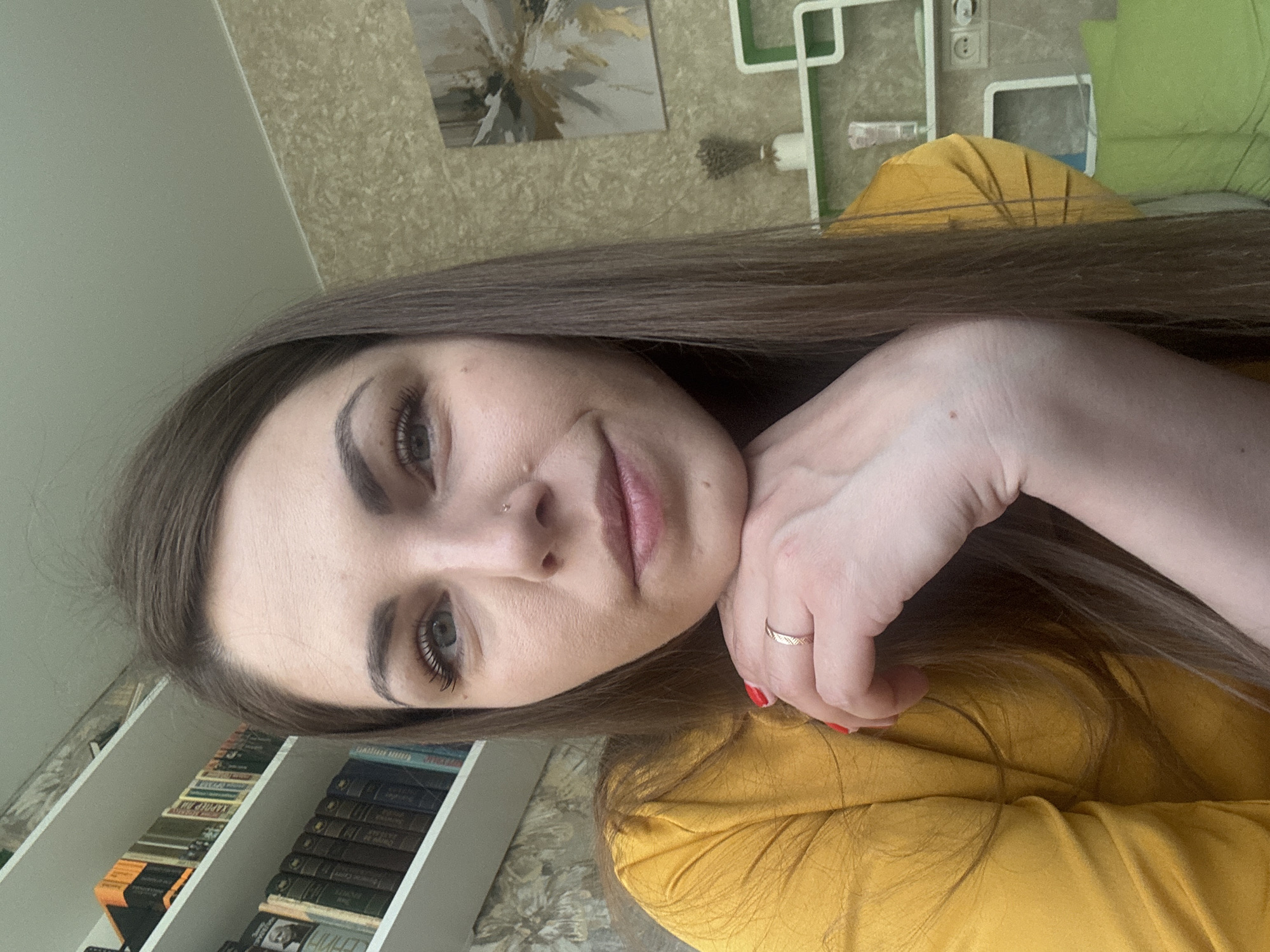 A woman with long brown hair is in an indoor setting. She is wearing a yellow shirt. She has a ring on her finger. A bookshelf filled with books is visible. The location is Раздолье, Russia.