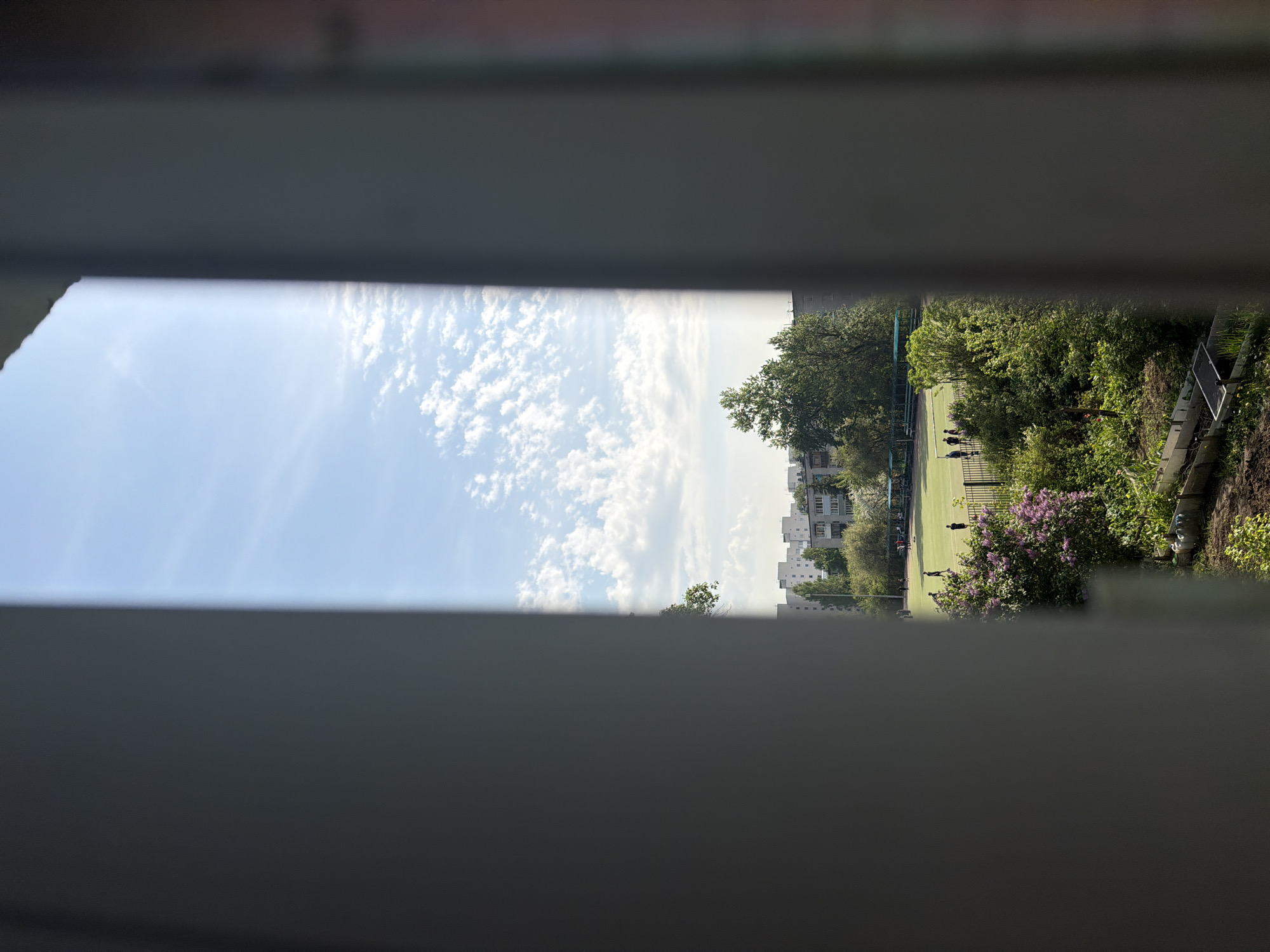 A view of a grassy field with a building visible beyond it is seen through a set of blinds. People are visible on the field. The sky is blue with clouds. There are trees and bushes to the right of the field.
It is not possible to identify a specific location or the city of Tokkari-Lend, Russia, from this image.