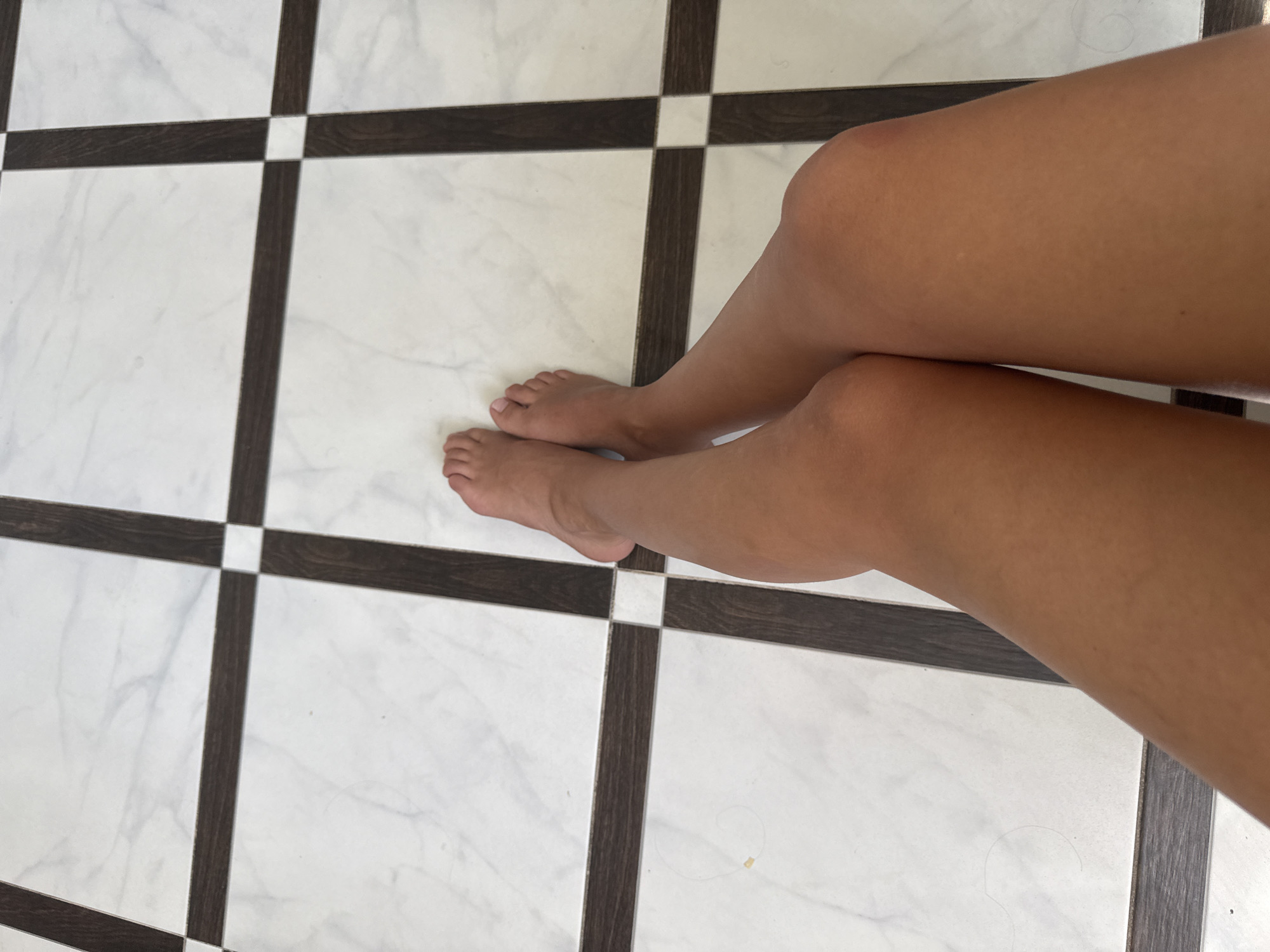 The image shows a close-up, overhead view of a person's legs and feet resting on a patterned floor. The floor is made of white marble-like tiles with dark brown rectangular accents forming a grid pattern. The person's legs are crossed at the knees, and their bare feet are visible. The scene appears to be indoors, possibly in a home or a public space with a tiled floor. There is no visible indication of the city of Chișinău, Moldova.