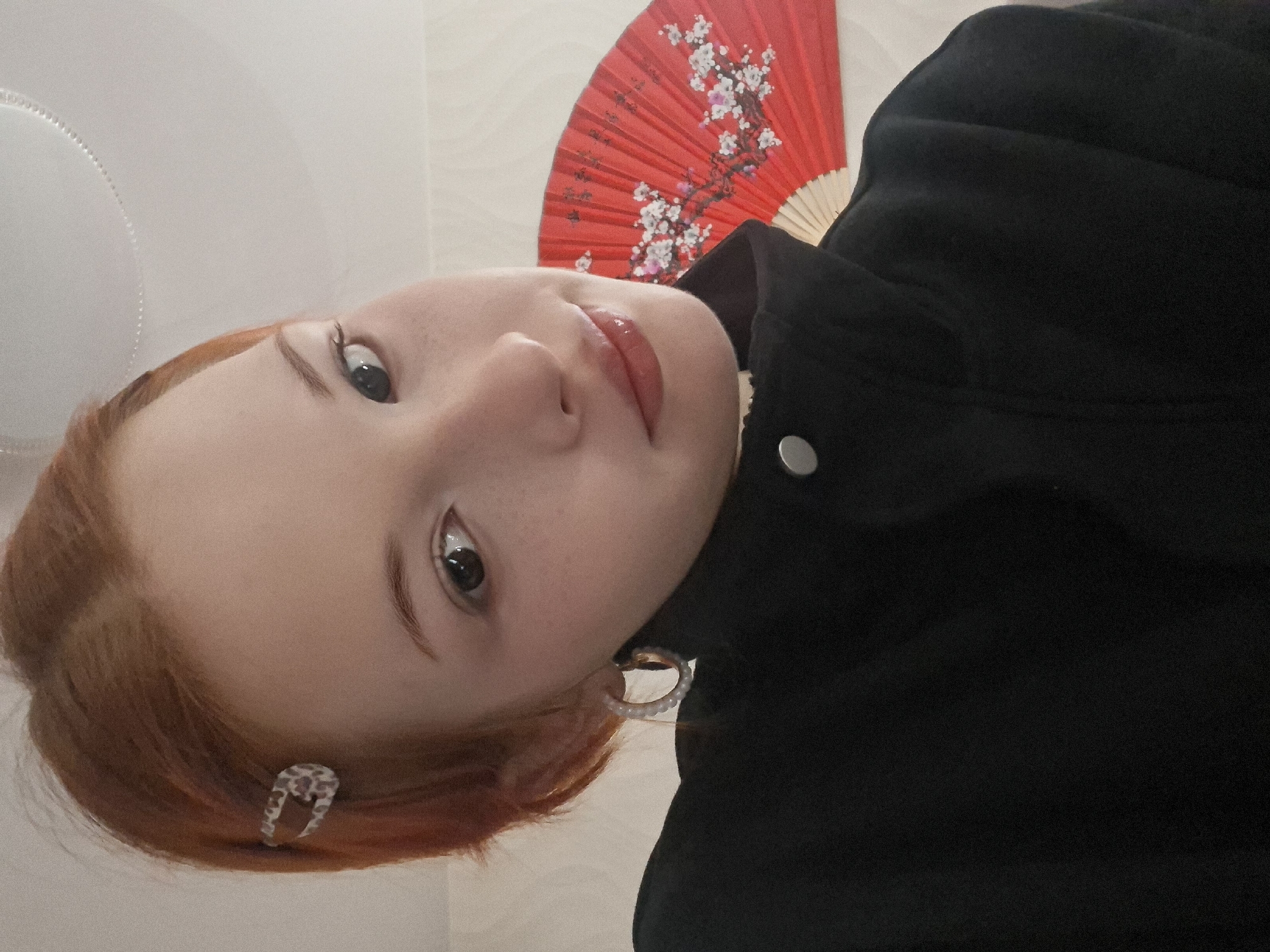 The image features a young woman with reddish-brown hair, seen from an upward angle with her head tilted to the right. She is looking directly at the camera with dark, open eyes and a subtle, soft expression, her lips glossy with a light pink color.
She is wearing a black jacket or top, with a silver button visible on the collar area. In her right ear, a hoop earring with small white beads or pearls is visible, and on the left side of her head, a silver-toned hair clip with a patterned design secures some of her hair.
Behind her head, a vibrant red hand fan is displayed. The fan features an intricate design of white and pink cherry blossoms (sakura) on branches. The background appears to be a light-colored wall or surface, suggesting an indoor setting. There is no visible text in the image. The location context provided is Chaikovsky, Russia.