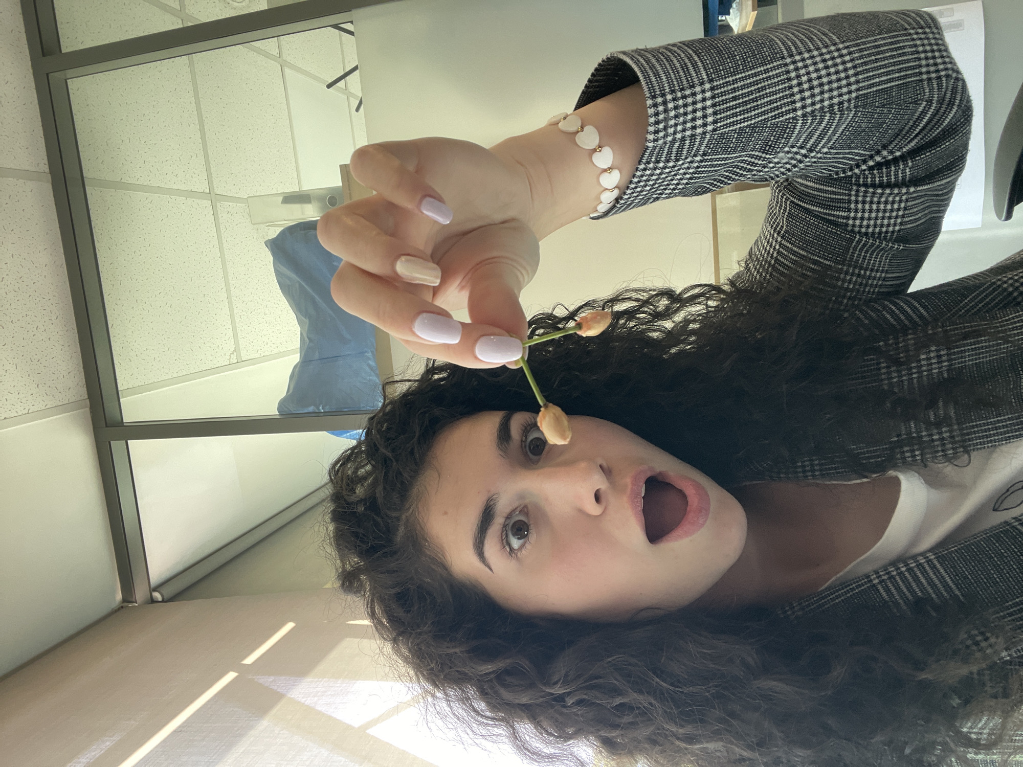 A young woman with long curly brown hair is holding a flower branch with three buds near her open mouth. She is wearing a white t-shirt and a gray and white plaid blazer. Her left wrist has a white bracelet with heart-shaped beads. Her nails are painted in light shades of pink and white. The location appears to be indoors, as there's a ceiling grid, window, and a blurred light blue object. There is no discernible information in the image to suggest it was taken in Sofia, Bulgaria.
