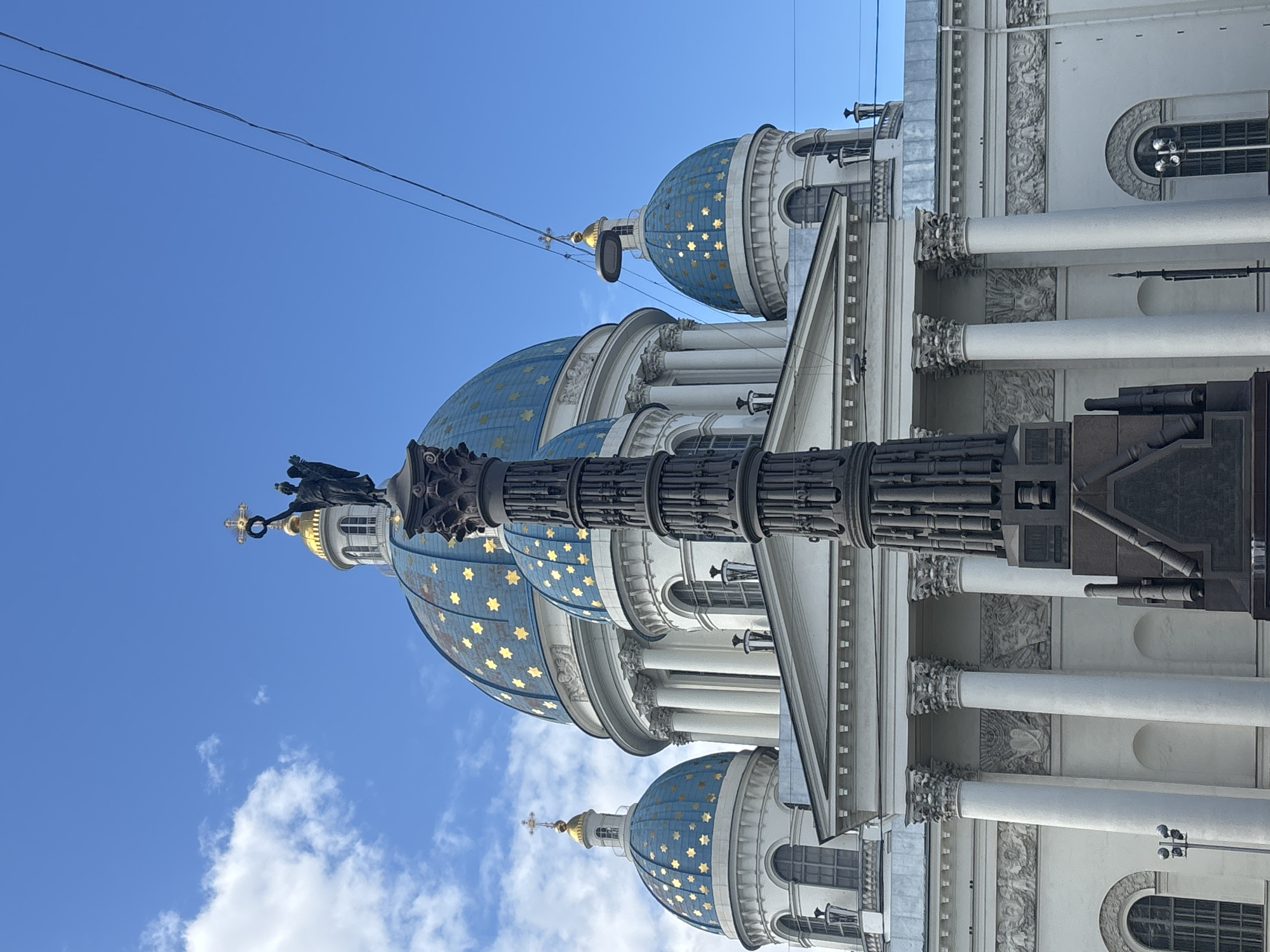 This is a low-angle, eye-level, full shot of the Trinity Cathedral in Saint Petersburg, Russia, under a bright blue sky with scattered clouds. The cathedral features multiple blue domes adorned with gold stars. A tall, dark monument with intricate carvings stands in the foreground, partially obscuring the cathedral. Overhead, power lines stretch across the frame. The cathedral's facade is white, with large columns and arched windows. The overall scene depicts an architectural landmark on a clear day.