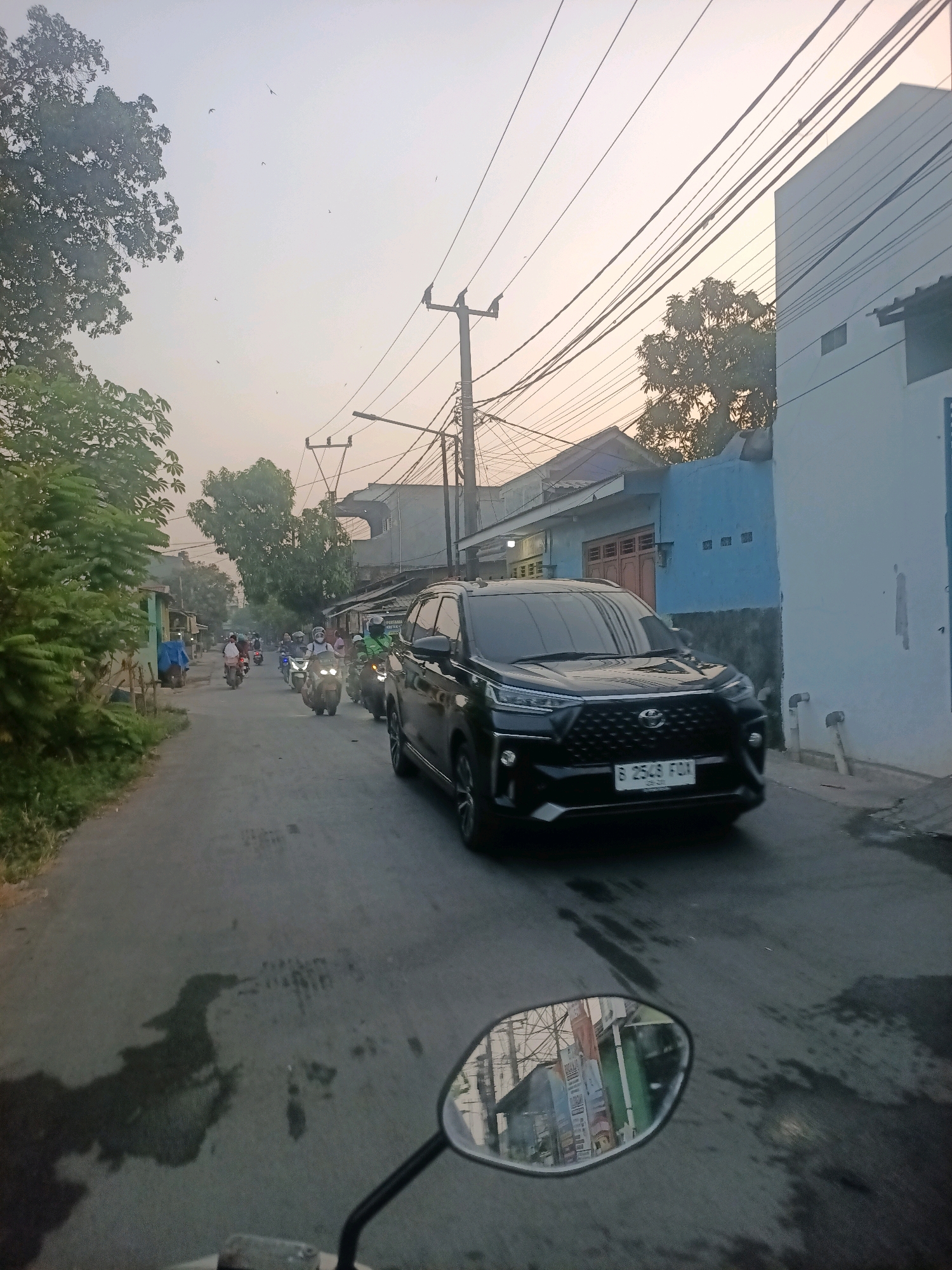 The image depicts a street scene in Wanajaya, Indonesia, likely during the early morning or late afternoon, given the soft, diffused light and the hazy sky. The perspective is from someone riding a motorcycle, evidenced by the motorcycle's rearview mirror prominently in the foreground, reflecting buildings and overhead wires. The street is paved and appears somewhat damp, with dark patches suggesting water or oil stains.
The street is lined with buildings on both sides. On the left, there are low-rise structures, some with blue tarps, and dense green foliage. On the right, the buildings are taller, with a prominent white-painted structure and a blue-painted wall. Overhead, a complex network of electrical wires crisscrosses the sky, supported by utility poles.
A black SUV is the most prominent vehicle in the foreground, positioned on the right side of the road, facing away from the viewer. Its license plate, partially visible, reads "B 2548 FDN". Further down the street, a procession of motorcycles and their riders are visible, moving in the same direction as the SUV and the motorcycle from which the photo is taken. The riders are wearing helmets, and some are carrying passengers. The overall atmosphere suggests a typical urban street with ongoing traffic.