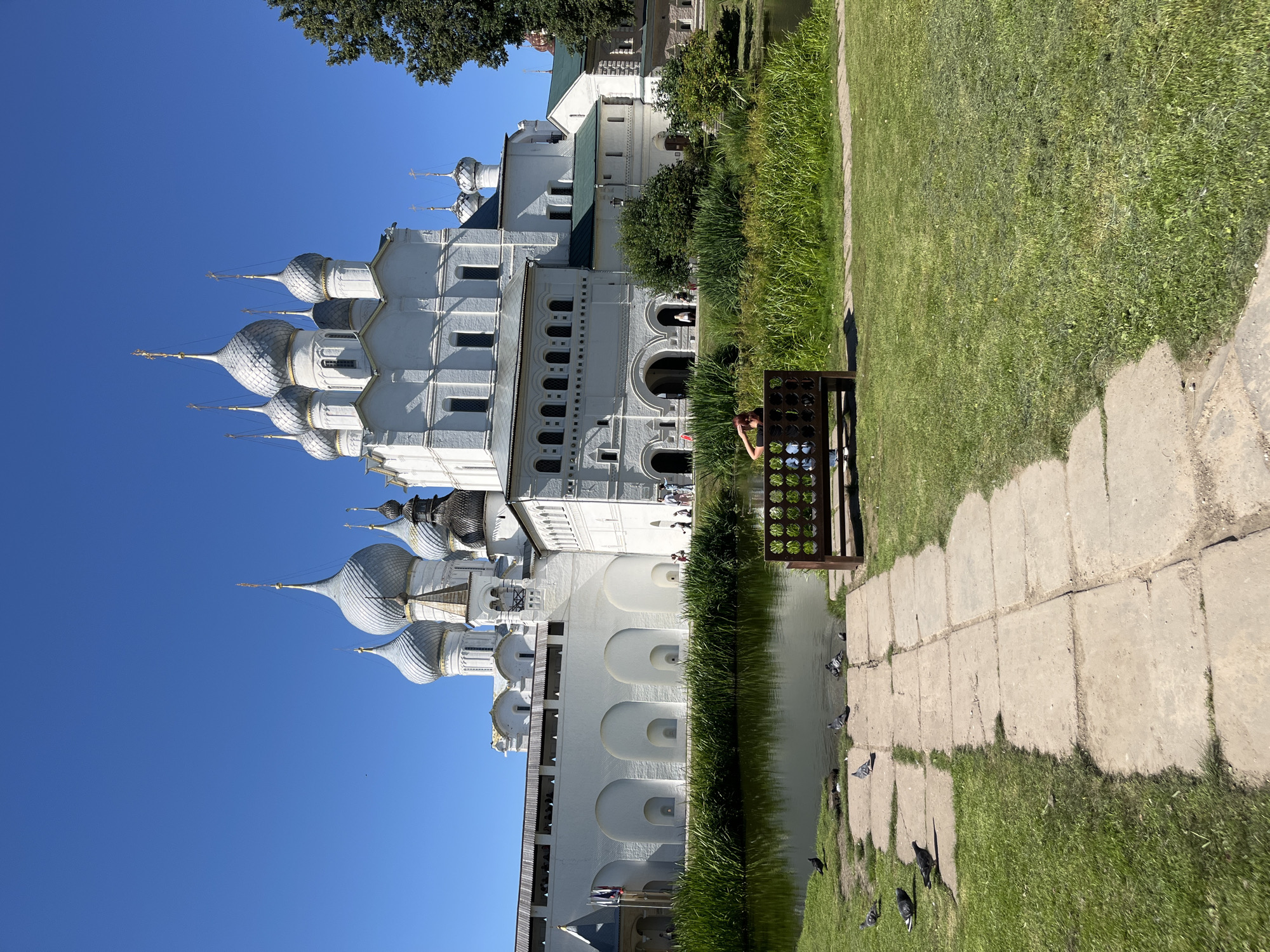The image shows a serene outdoor scene at the Rostov Kremlin in Novo-Nikolskoye, Russia. Dominating the background is the majestic white architecture of the kremlin, featuring multiple onion-shaped domes topped with golden crosses, reaching towards a clear blue sky. A long, white wall with arched alcoves and wooden eaves runs along the lower part of the kremlin structure, separating it from the foreground.
In the middle ground, a tranquil body of water reflects the sky and the surrounding greenery. Tall, lush green reeds and grass line the edges of the water. A lone woman sits on a wooden bench with an intricate circular design in the back, facing away from the camera and towards the water and kremlin. She appears to be looking out at the scenery.
The foreground is occupied by a paved pathway made of large, rectangular stone slabs, which leads from the lower right of the frame towards the water. This path is partially bordered by a neatly mown green lawn. Several pigeons are scattered on the path and near the water's edge, some pecking at the ground. The overall atmosphere is peaceful and picturesque, bathed in bright sunlight.