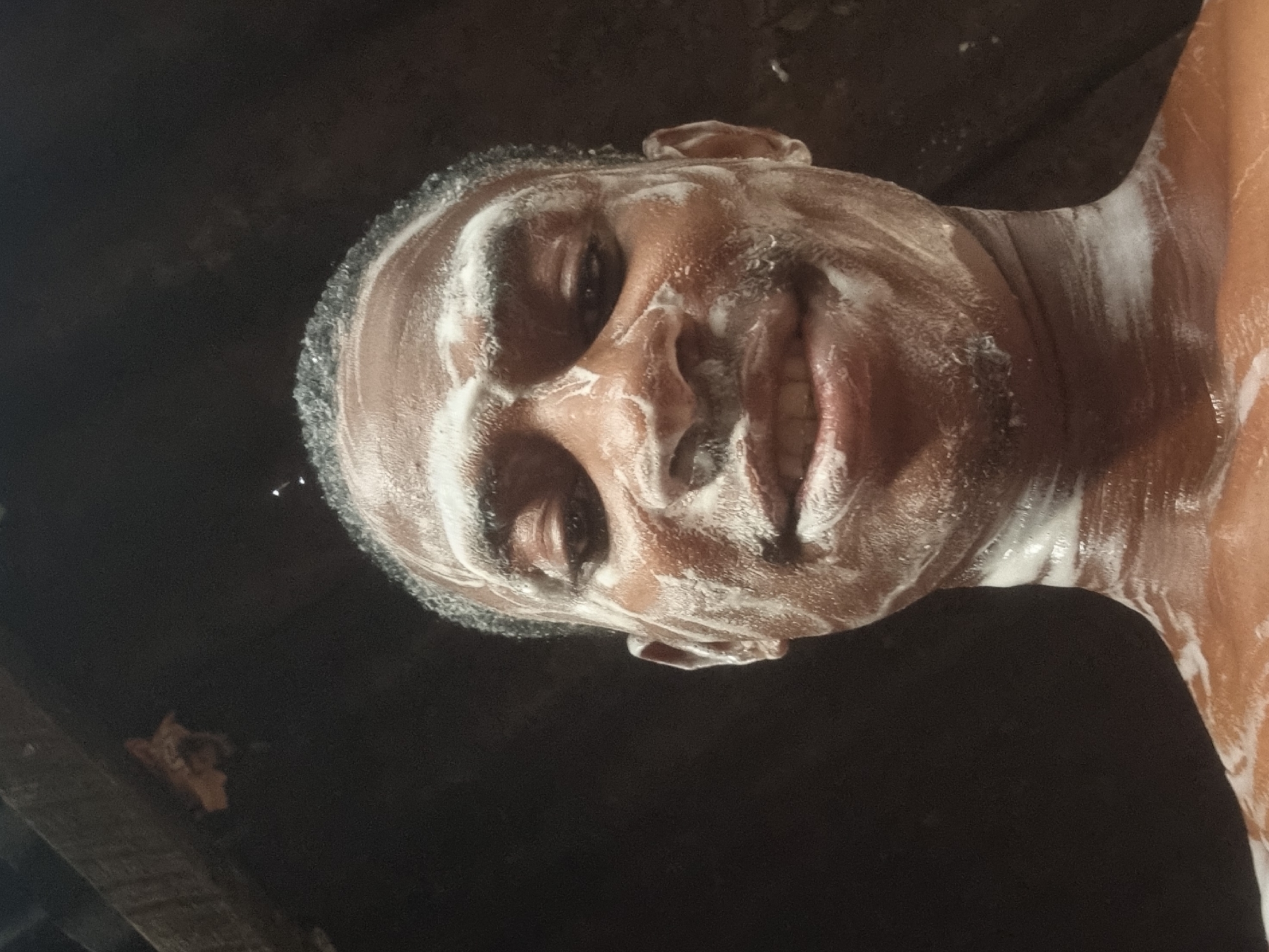A close-up, rotated portrait shows an individual with dark skin, likely male, covered in white, frothy suds on their face, hair, and upper torso. The person is looking directly at the viewer with a slight smile or smirk, indicating a relaxed or playful mood. Their short, dark hair is also extensively covered in the foam.
The scene suggests the person is in the process of washing or showering. The background is dark and indistinct, hinting at an indoor setting with low lighting, possibly a bathroom. The lighting emphasizes the contrast between the dark skin and the bright white foam. The image is oriented with the person's head on the left side of the frame, tilted upwards, and their shoulder extending to the right. This activity takes place in Onitsha, Nigeria. There is no visible text in the image.