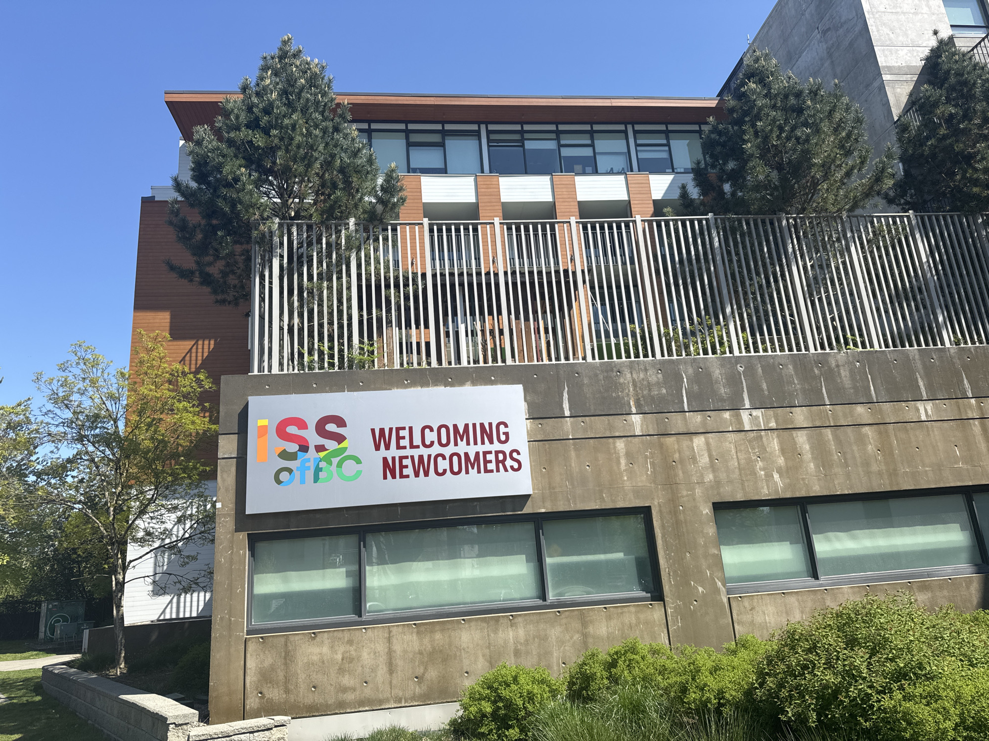 The image shows the Immigrant Services Society of British Columbia (ISSofBC) building. The foreground features a concrete retaining wall with a sign that reads "ISS of BC WELCOMING NEWCOMERS". The ISSofBC logo is multicolored. The building behind the wall has a combination of brown wood paneling and white facade elements. There are two trees on the upper level behind the retaining wall, partially obscuring the building. The sky is blue. The location is likely Vancouver, Canada, as ISSofBC is based there. There are also some other trees and shrubbery around the concrete retaining wall. The windows behind the sign on the concrete wall are tinted or covered. The fence behind the concrete wall is made of vertical silver bars.