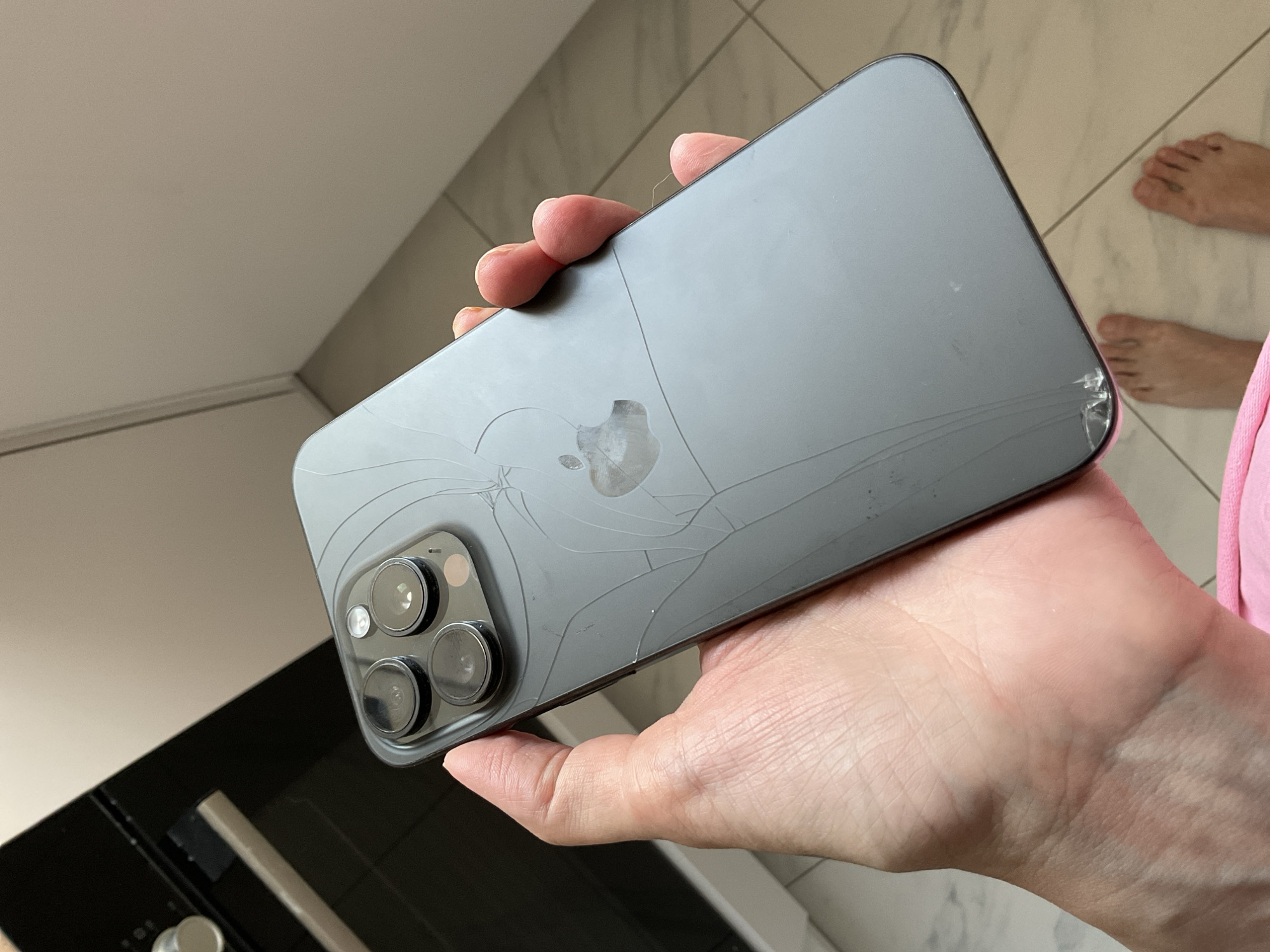A person holds a gray iPhone with a cracked back in their hand. The background includes a kitchen counter, tiled walls, a black appliance (likely an oven), and a tiled floor. A person's feet are visible in the upper right corner. It is not possible to determine the location as Saratov, Russia from this image alone.