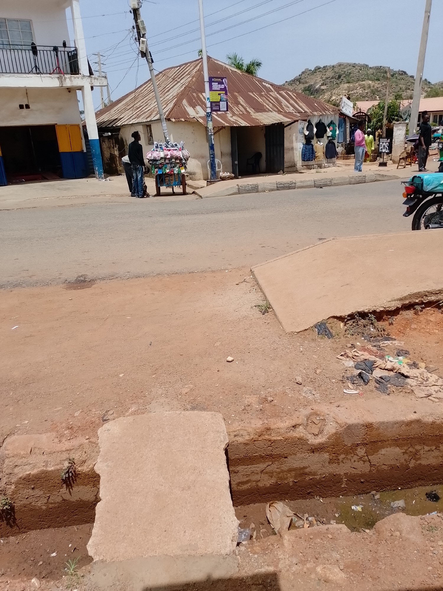 The scene is in Gura Top, Nigeria. A streetscape shows a road with a two-story building on the left side, painted white with blue and yellow accents. Across the road is a single-story structure with a corrugated metal roof, some of which appears to be rusted. People are seen on the street: two figures near a cart covered with what appear to be mobile phone accessories, one in a pink shirt standing near clothing displayed outside the single-story structure, and others at a distance. A motorcycle is parked on the right side of the road. The foreground contains an exposed drainage ditch, partially covered with a concrete slab, with visible trash and stagnant water. The background features hills in the distance. The sky is visible and appears clear.