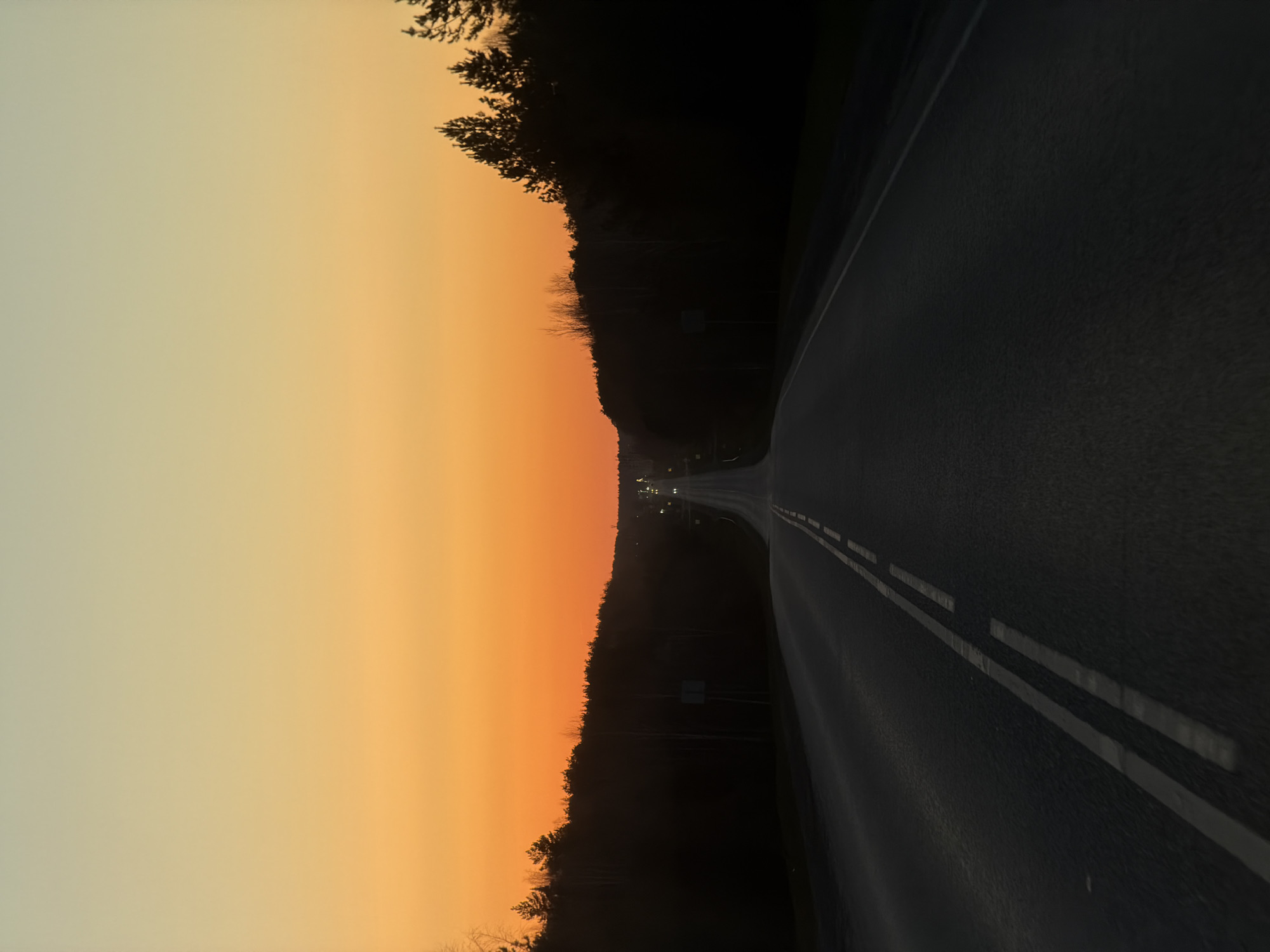 The image depicts an asphalt road with double white lines running along the center. The road curves slightly to the right, leading away into the distance. On the left side of the road is a dense, dark tree line with a gradient of orange and pale yellow sky above it. The sky suggests either a sunrise or sunset. In the distance, faint lights indicate a possible town or populated area, suggesting the image may have been taken near a city. Due to the presence of trees, lights in the distance, a curving road, and the gradient in the sky the location of this image could be in the city of Chaikovsky, Russia.