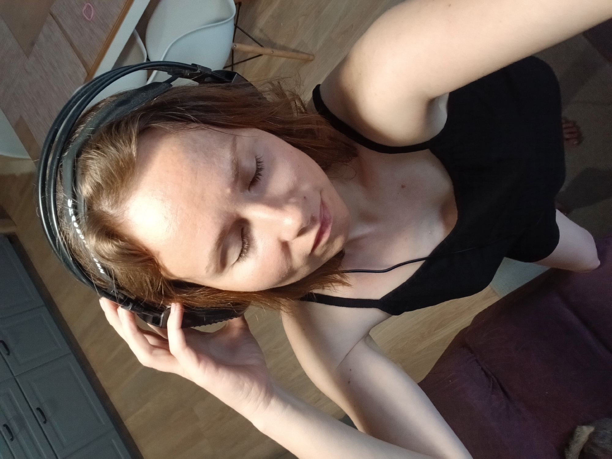 The image shows a person with light brown hair wearing large black headphones. The person's eyes are closed, and their head is tilted back slightly. They are wearing a black tank top. The person is holding one of the headphone cups with their left hand. The background consists of a wooden floor and a glimpse of a grey cabinet with handles. A purple sofa is visible in the lower right corner. The lighting suggests it may be daytime, possibly indoors with natural light. The setting appears to be a home environment.