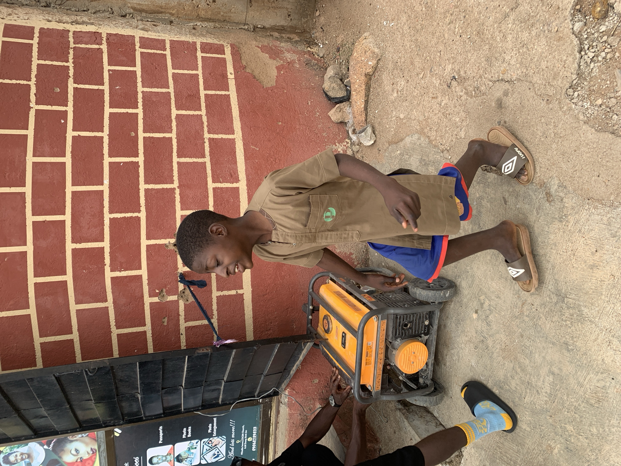 The image shows a young person in Jos, Nigeria, standing beside a portable generator, likely a common sight given the frequent power outages in the city. The youth is wearing a khaki-colored shirt and blue shorts with red trim, and brown flip-flops with "LUOFU" written on them. The generator is yellow and black, and someone wearing a black shirt, and blue socks and a black shoe is seen working with the generator.
Behind the youth is a wall painted with a faux brick pattern in red and tan. To the left is a section of a storefront with a sign advertising services like passports, photo books, and photo enlargements. The sign includes the phrase "And lots more!!!" and the contact information for a business named "Dodin howo, COCIN Church, A.M Dung Street Jos, 09068789823." The ground is a mix of concrete and dirt. The scene is illuminated by natural light.
