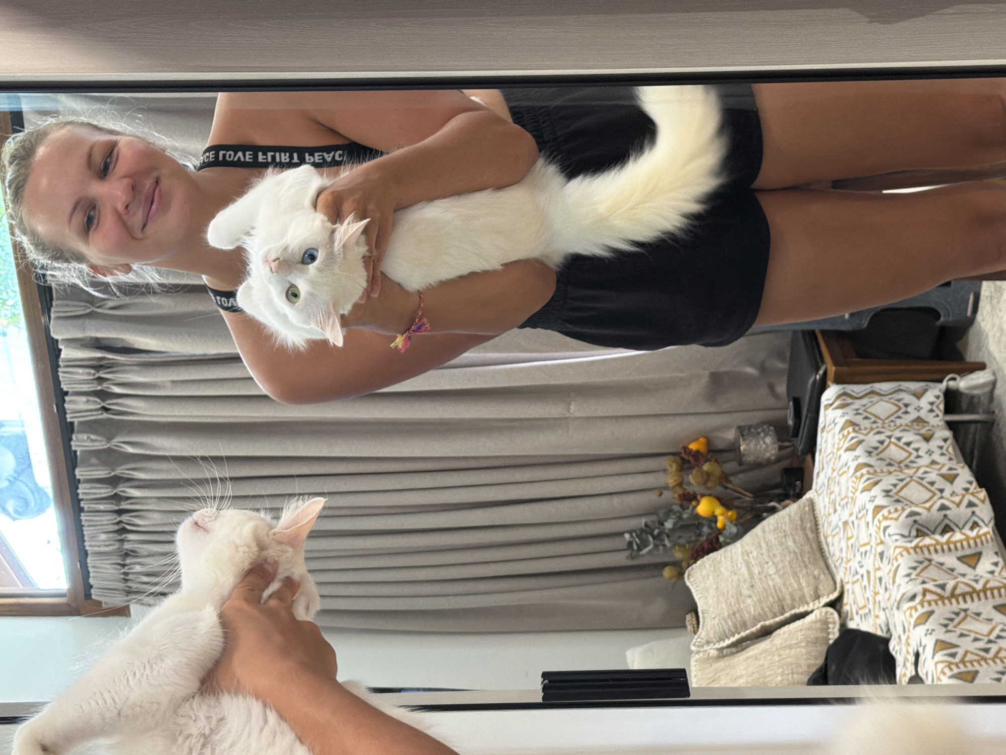 The image shows a reflection in a mirror. A fair-skinned woman is holding a white cat with heterochromatic eyes (one blue, one green). The woman is wearing a black sports bra and black shorts. A second white cat is visible being petted. The background includes gray curtains, a decorative vase with dried flowers, and a patterned bedspread. The location is inside an RV, city of Unknown, Unknown.