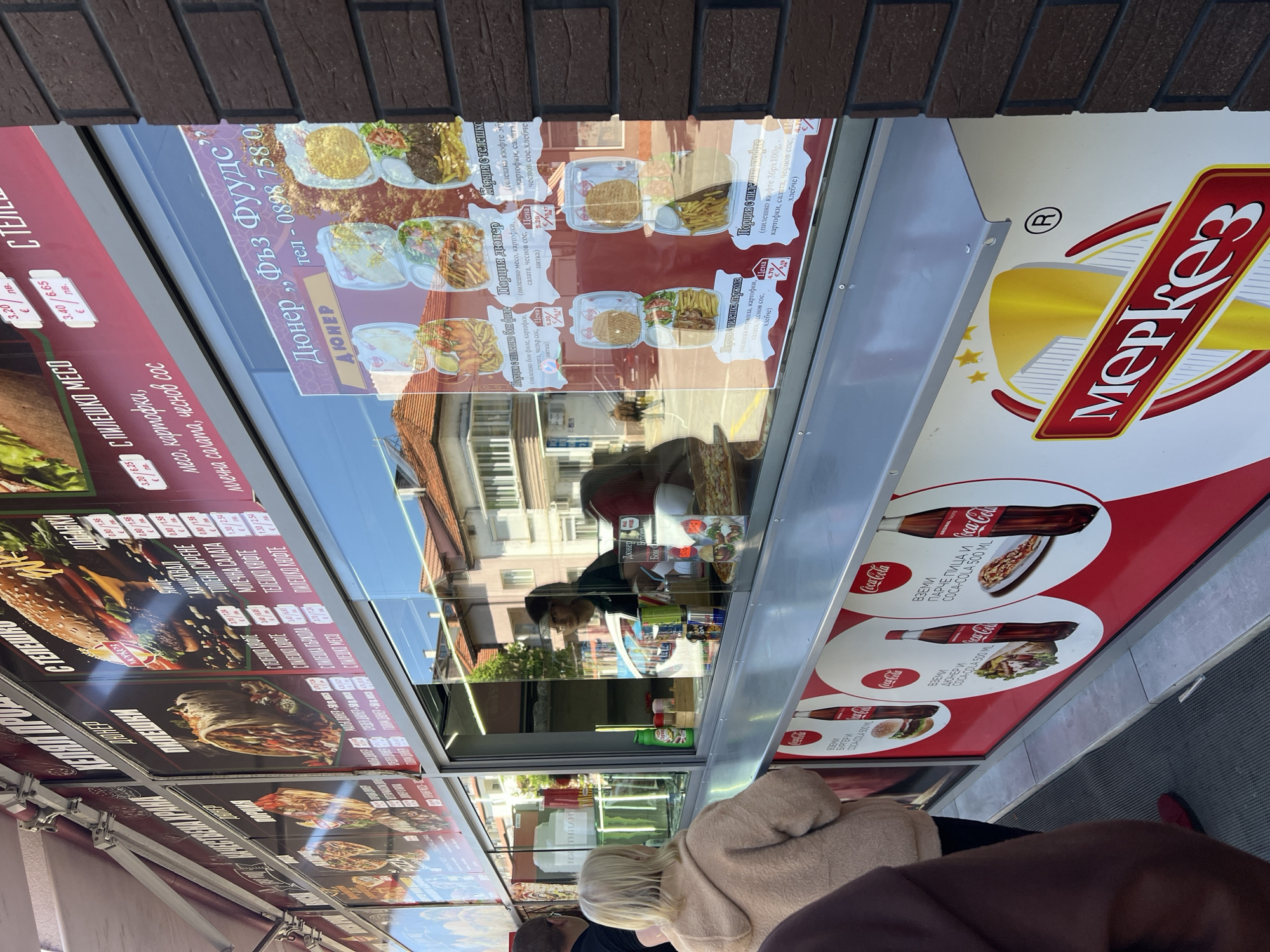 The image captures a street scene in Gotse Delchev, Bulgaria, focusing on a food stall. The stall displays menus with images and prices of various food items like doner kebab, burgers, and meat dishes. Brand advertising for "Merkess" and Coca-Cola is also visible on the stall's exterior.
Several people are visible, primarily customers standing in front of the stall. One customer with blonde hair is wearing a beige coat, while another person in the foreground is wearing a dark brown jacket. Through the glass of the stall, the food preparer is partially visible, wearing a dark shirt and glasses.
The environment suggests an urban setting, with buildings visible in the background and brickwork on the stall's side. The overall scene depicts a typical street food vending operation with customers interacting and waiting to order.