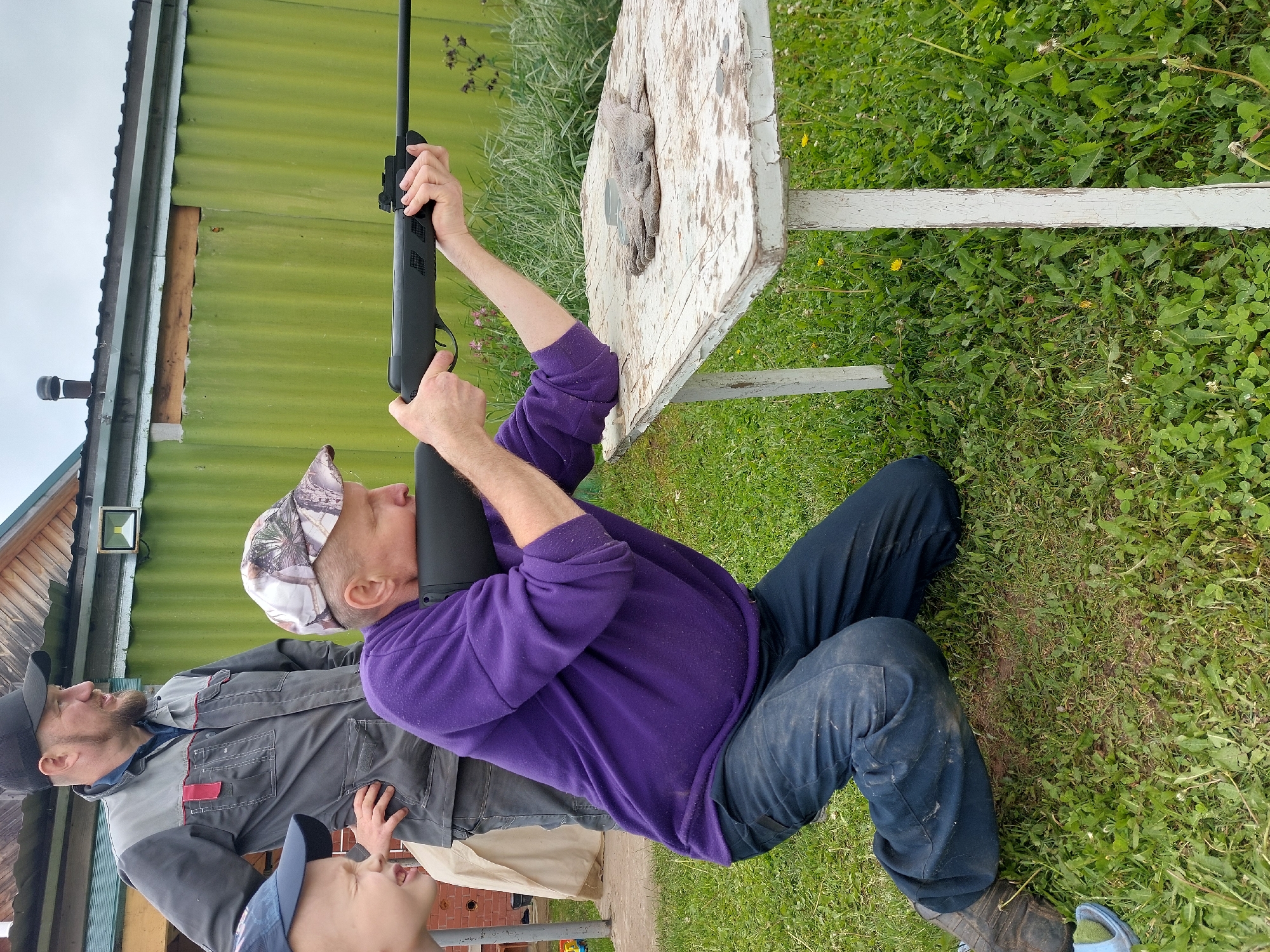 In Большой Букор, Russia, an outdoor scene depicts three males gathered around a shooting activity on an overcast day. A man, appearing middle-aged to older, kneels on his right knee in the foreground, wearing a camouflage baseball cap, a purple long-sleeved top, and dark blue trousers. He is aiming a black air rifle, resting the rifle's fore-end on a small, rectangular white wooden table with significantly peeling paint. His right hand is on the rifle's grip with a finger near the trigger, and his left forearm supports the rifle on the table. A pair of worn brown work gloves rests on the right side of the table.
To the left and slightly behind the kneeling man, another man, appearing adult, stands upright. He wears a black baseball cap, a grey and black work jacket with red detailing, and dark trousers, observing the kneeling man with his hands on his hips. Partially visible on the far left, a young boy with a light complexion and a blue baseball cap looks towards the rifle with an open-mouthed expression. A light blue sandal is visible on the grass in the bottom-left corner.
The ground is covered in green grass mixed with dandelions and clover. In the background, a green corrugated metal structure extends across the mid-ground, featuring a section of exposed wood near its top. Above this, a wooden building with a dark roof and a dark pipe-like chimney is visible. A wall-mounted outdoor light fixture is attached to the green structure on the right side. The sky is a uniform light grey, indicating an overcast condition.