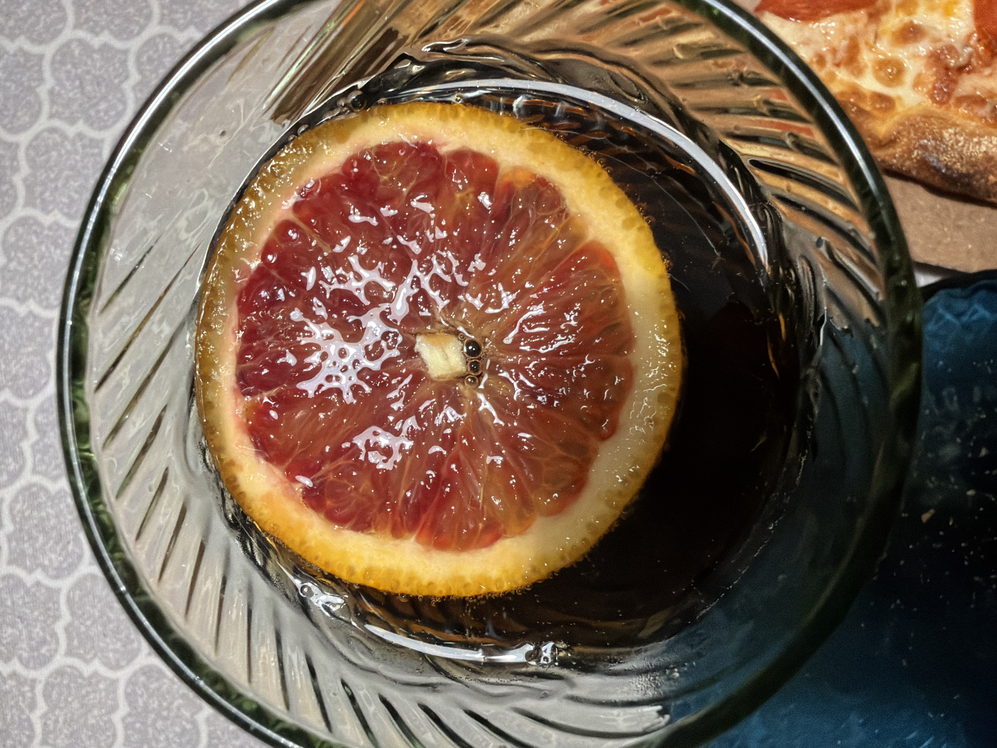 A close-up, overhead shot shows a glass containing a dark liquid and a slice of blood orange. The glass is clear with vertical ridges on the exterior. The blood orange slice floats on top of the liquid. A portion of a pizza crust with toppings is visible in the upper right corner. The glass sits on a surface with a patterned design. The location is unknown, and there is no evidence to indicate it is in Votkinsk, Russia. No people, actions, or interactions are depicted.