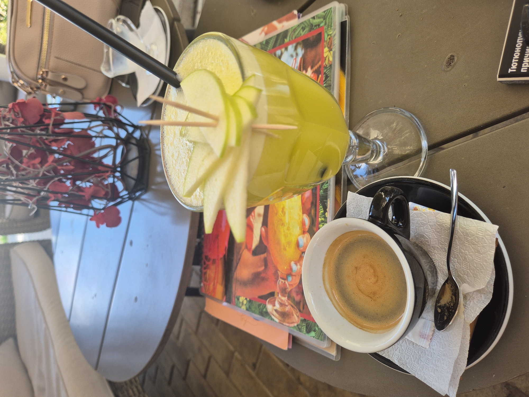 The image shows a close-up of a table at an outdoor cafe. On the table, there is a glass of yellow juice with apple slices and toothpicks, a demitasse cup of espresso with crema, a spoon, a napkin, and a saucer. In the background, there is a purse and a decorative object with red flowers. The setting appears to be a relaxing outdoor area. The table surface is a speckled brown color. The context suggests a leisurely moment at a cafe.