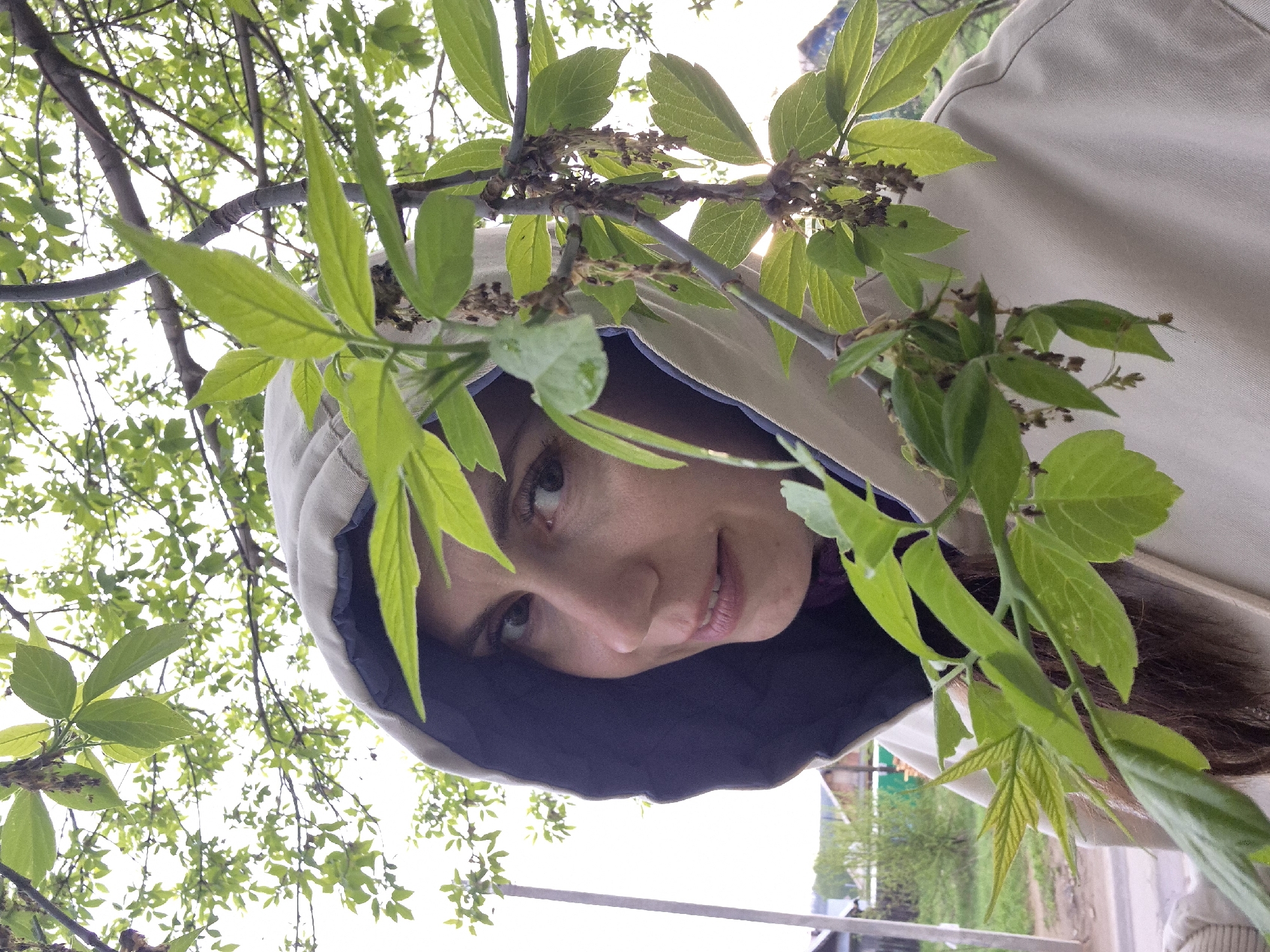 A woman with brown hair is wearing a hooded beige jacket and is surrounded by green leaves and branches. She is looking towards the camera with a slight smile. The hood of her jacket is lined with dark blue fabric. In the background, there is greenery, trees, and a glimpse of buildings and structures suggesting an outdoor environment possibly in a park or residential area.