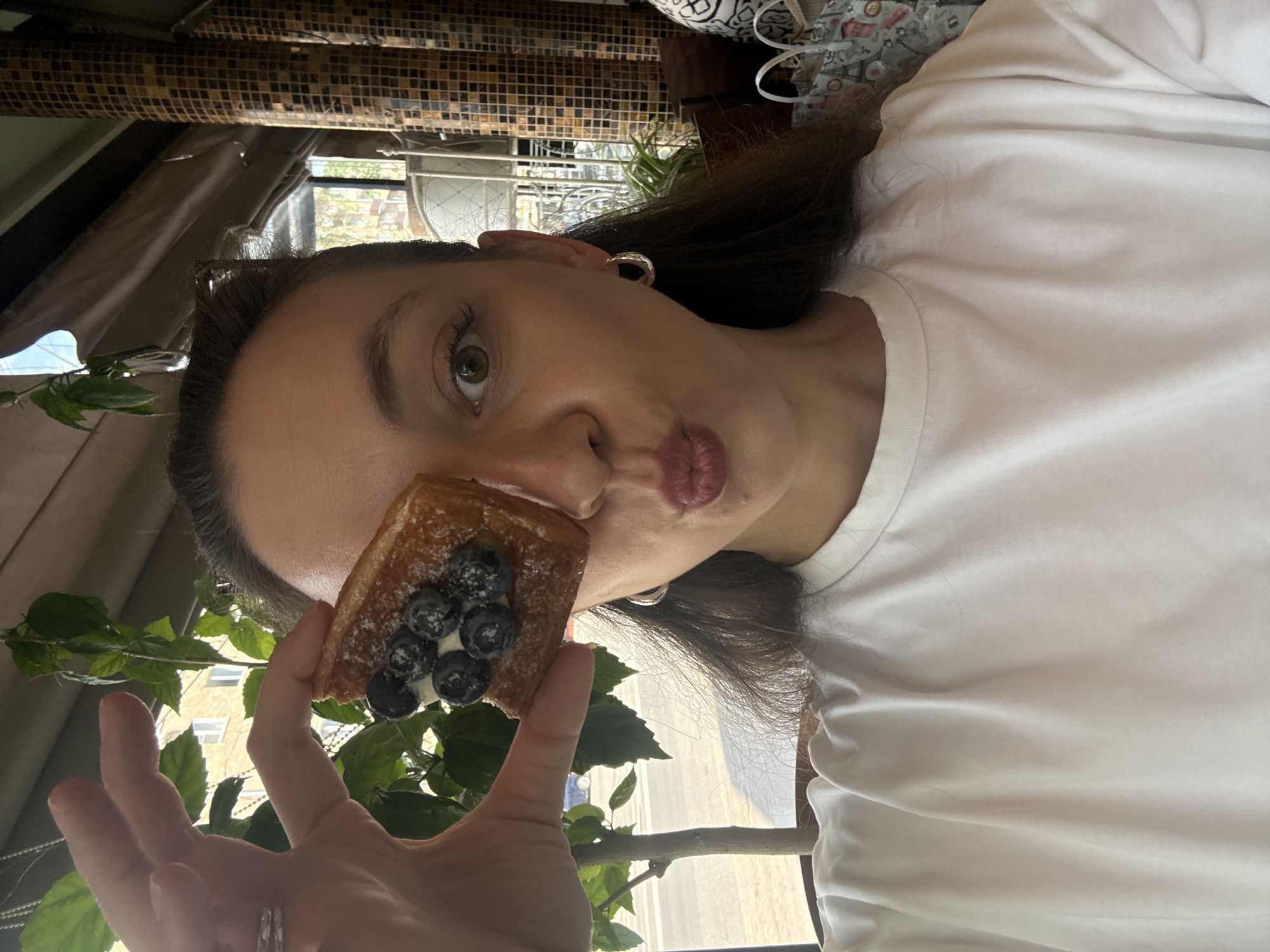The photo shows a woman in a white t-shirt holding a pastry topped with blueberries in front of her face, with a building visible through the window behind her. The location is Moscow, Russia.