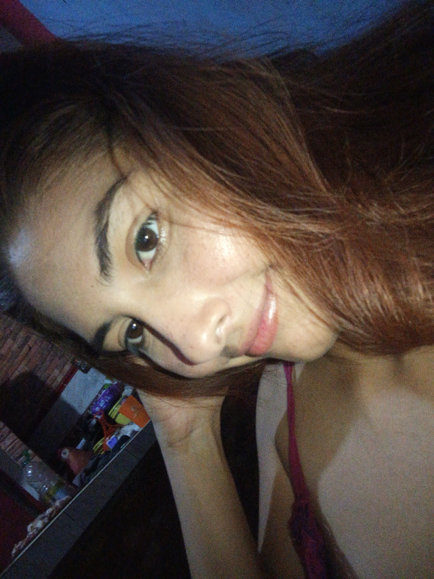 A close-up photograph shows a person with light brown hair and brown eyes, with slightly freckled skin. The person is slightly angled towards the viewer and appears to be smiling faintly. A thin strap, possibly from a garment, is visible at the neck area. In the background, there is a shelf with various items, including bottles and packaged goods, and a wall made of bricks is behind the shelf. The setting may be indoors. The specific location in Juan Griego, Venezuela, is not identifiable from the visual information in the image.