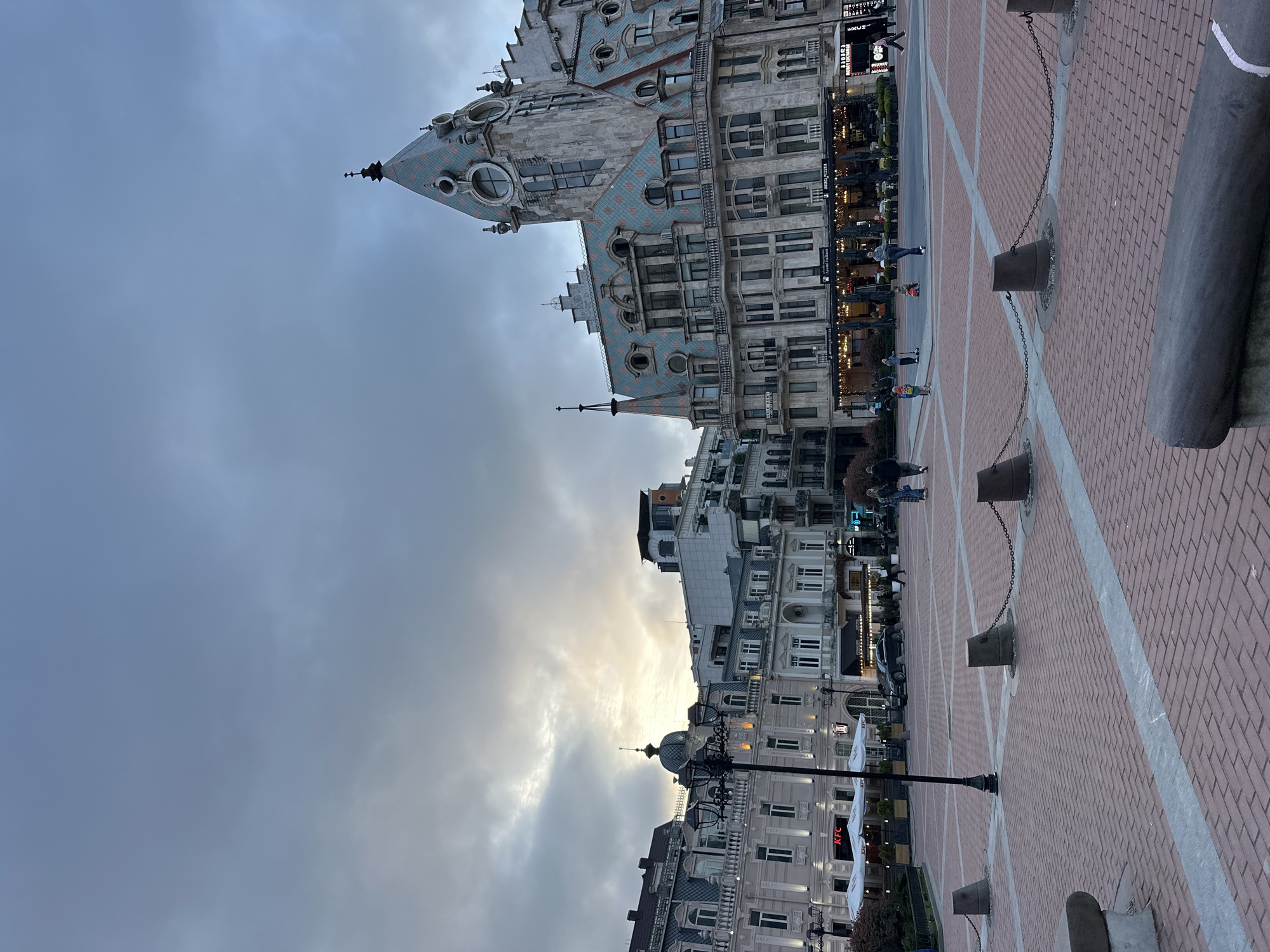 The image displays an urban plaza or square, likely in a city, under an overcast sky with a bright patch near the horizon. The scene appears to be taken during daylight hours, possibly late afternoon or early morning, given the soft, diffused light and the visible break in the clouds.
On the left side of the frame stands a striking, multi-story building with ornate architecture. Its facade is light-colored, possibly stone or stucco, and features numerous windows. The most prominent characteristic is its complex roofline, which is covered in a distinctive pattern of blue-green and reddish-orange tiles. The roof includes multiple gables, circular windows or decorative elements, and several slender spires that pierce the sky.
To the right and extending into the background, other multi-story city buildings line the perimeter of the square. These buildings also exhibit classical architectural elements, such as arched windows and decorative cornices. Some appear to house commercial establishments on their ground floors, indicated by storefronts and outdoor seating areas with white umbrellas or awnings. A red "KFC" logo is clearly visible on an awning of one such establishment in the lower right background.
The foreground and midground of the image are dominated by a large, paved public space. The paving consists primarily of reddish-brown brick laid in a patterned arrangement, interspersed with light grey stone stripes that form a grid across the plaza. Along the right side of this paved area, a series of dark, sturdy bollards or posts are installed, connected by chains, creating a decorative or functional barrier. A tall, black street lamp with multiple fixtures is also visible in the lower right portion of the plaza.
Several people are scattered throughout the square, walking or gathered near the buildings, suggesting typical daily activity in a public space. Their specific actions or features are not distinctly clear due to the distance from the camera. The sky above is largely covered by grey clouds, but a luminous area near the horizon, suggesting the position of the sun, creates a subtle glow.