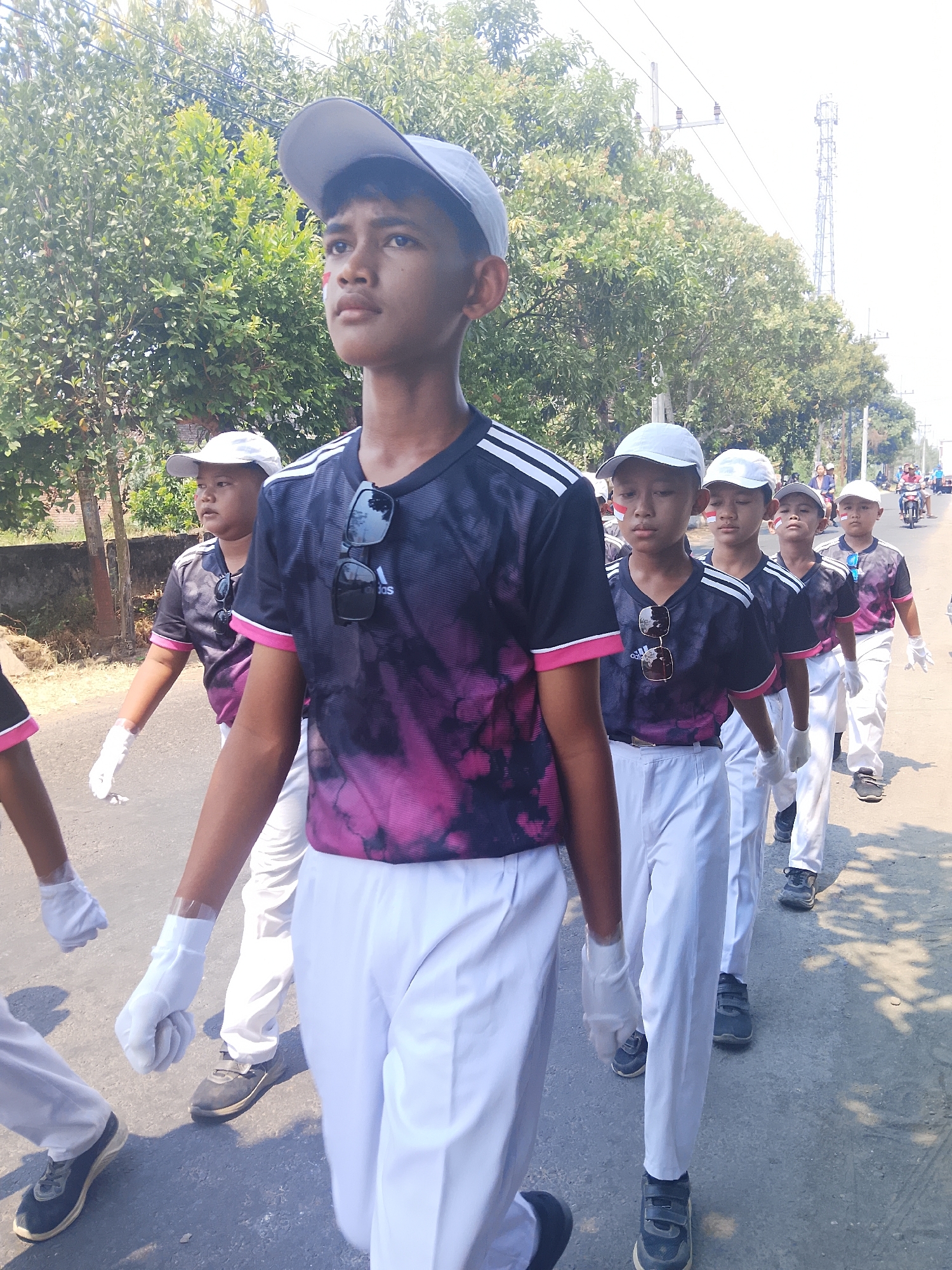 The image shows a group of young Indonesian boys marching in what appears to be a parade or procession on a sunny day in Bangsri, Indonesia. The boys are wearing matching dark t-shirts with a pink and black tie-dye pattern, white pants, white gloves, and white baseball caps. Some of the boys have red and white face paint on their cheeks, possibly representing the Indonesian flag. They are walking on a paved road lined with trees and a low brick wall on the left side of the image. In the background, there are power lines, a telecommunications tower, and other people and vehicles, including a motorcycle. The overall scene depicts a community event or school activity.