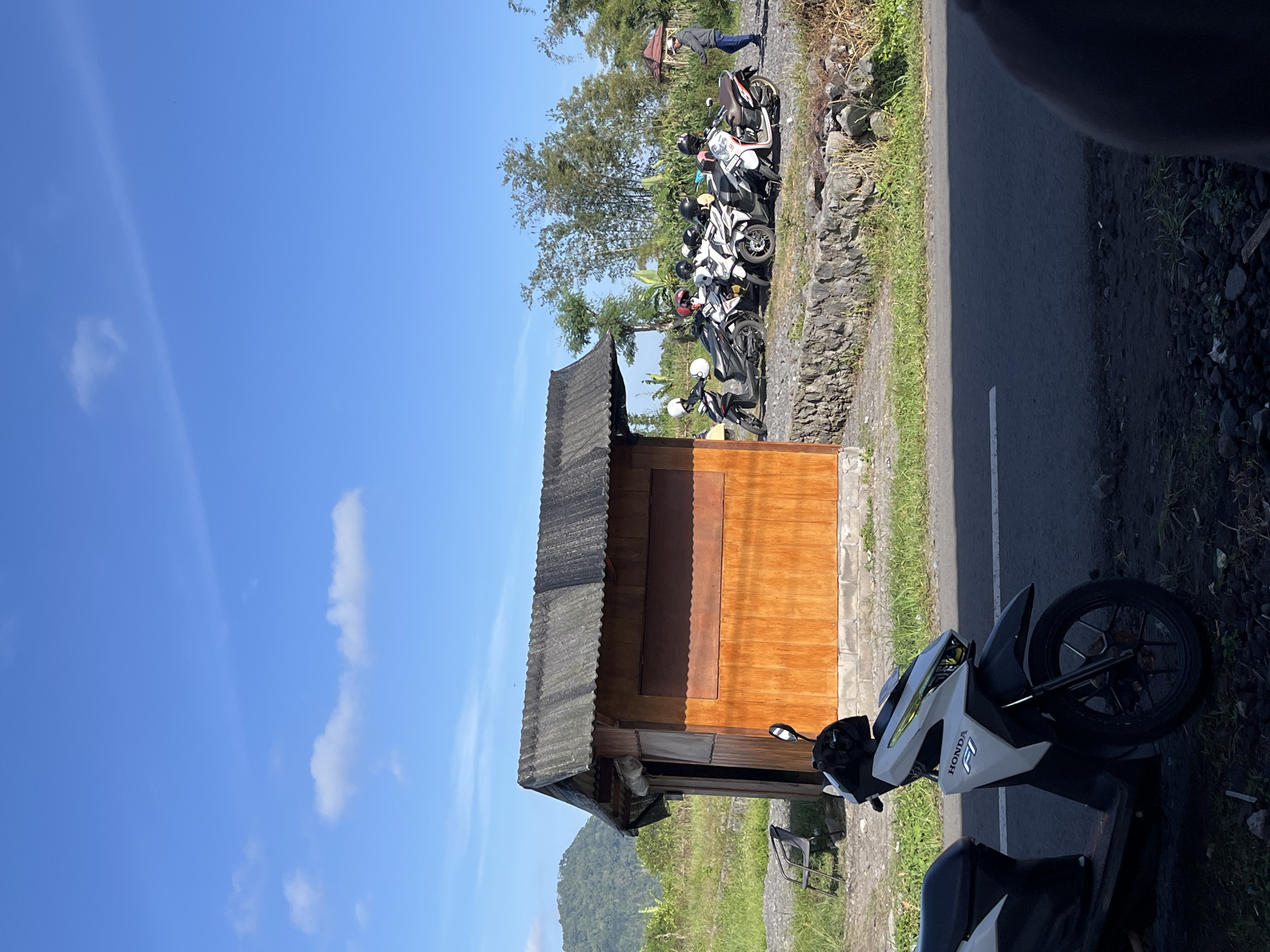 The image shows a scene on a sunny day in Oro-oro Ombo, Indonesia. In the foreground, a white and black Honda F1 scooter is parked on a gravel surface next to an asphalt road. The scooter has a black seat and handlebars. Behind the scooter, a small wooden shack with a corrugated metal roof stands. The shack has an open doorway on the left side and a large, dark wooden panel on its front. To the right of the shack, a line of several motorbikes, mostly white and black with helmets on their seats, are parked. One person wearing a grey jacket and blue jeans is standing near the motorbikes, facing away from the viewer. In the background, a green, tree-covered hill is visible under a bright blue sky with scattered white clouds. The environment is rural, with grass and stones visible around the shack and road.