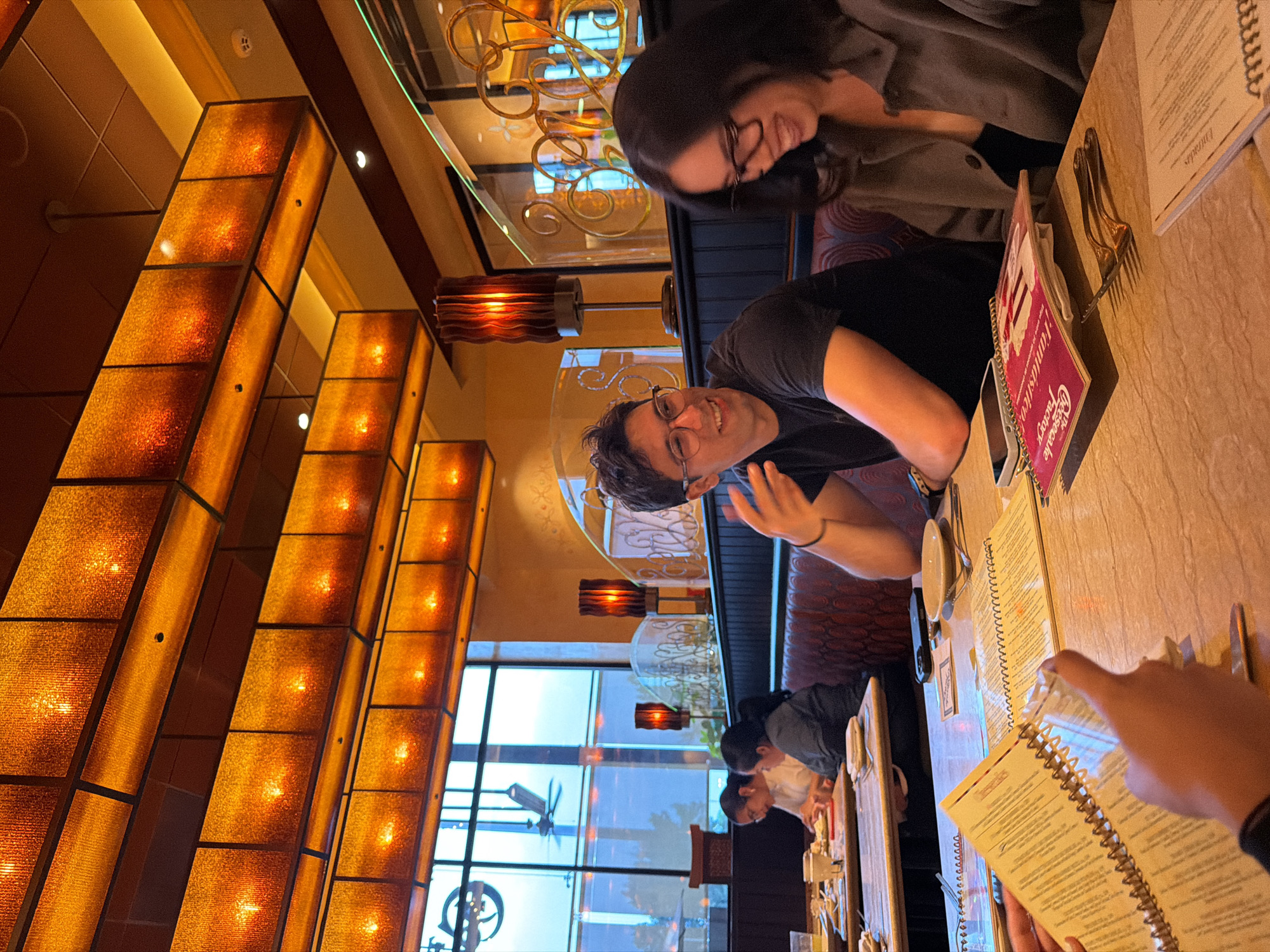 This media file captures an indoor dining scene at what appears to be a Cheesecake Factory restaurant in Puebla, Mexico. The image is taken from a slightly elevated perspective and is rotated 90 degrees clockwise, showing a warm and inviting ambiance with a focus on people and decorative lighting.
In the mid-ground, two individuals are seated at a light-colored table. One person, a male wearing glasses and a black t-shirt, is looking directly at the camera with a friendly smile, his left hand slightly raised. Opposite him, a female with long dark hair and glasses, dressed in a grey top, is leaning forward and looking down, possibly at a menu or engaged in conversation. On the table in front of them are menus, including a prominent red spiral-bound menu clearly displaying "The Cheesecake Factory" logo and name. Several pieces of silverware, specifically forks, are also visible on the table.
The background features large windows, indicating that it is likely late afternoon or early evening, with natural light still visible but interior lights already on. Through the windows, blurred outdoor elements like buildings and possibly a distant ceiling fan can be seen. The left side of the frame is dominated by a wall or partition adorned with numerous large, vertical rectangular light fixtures, featuring amber-toned, textured panels that emit a warm, golden glow. Smaller decorative pendant lights with reddish-brown shades are also suspended throughout the space. The ceiling reflects a decorative, ornate chandelier-like fixture. Further back in the restaurant, other patrons are visible seated at tables. The overall atmosphere appears relaxed and social, with the subjects engaged in their dining experience.