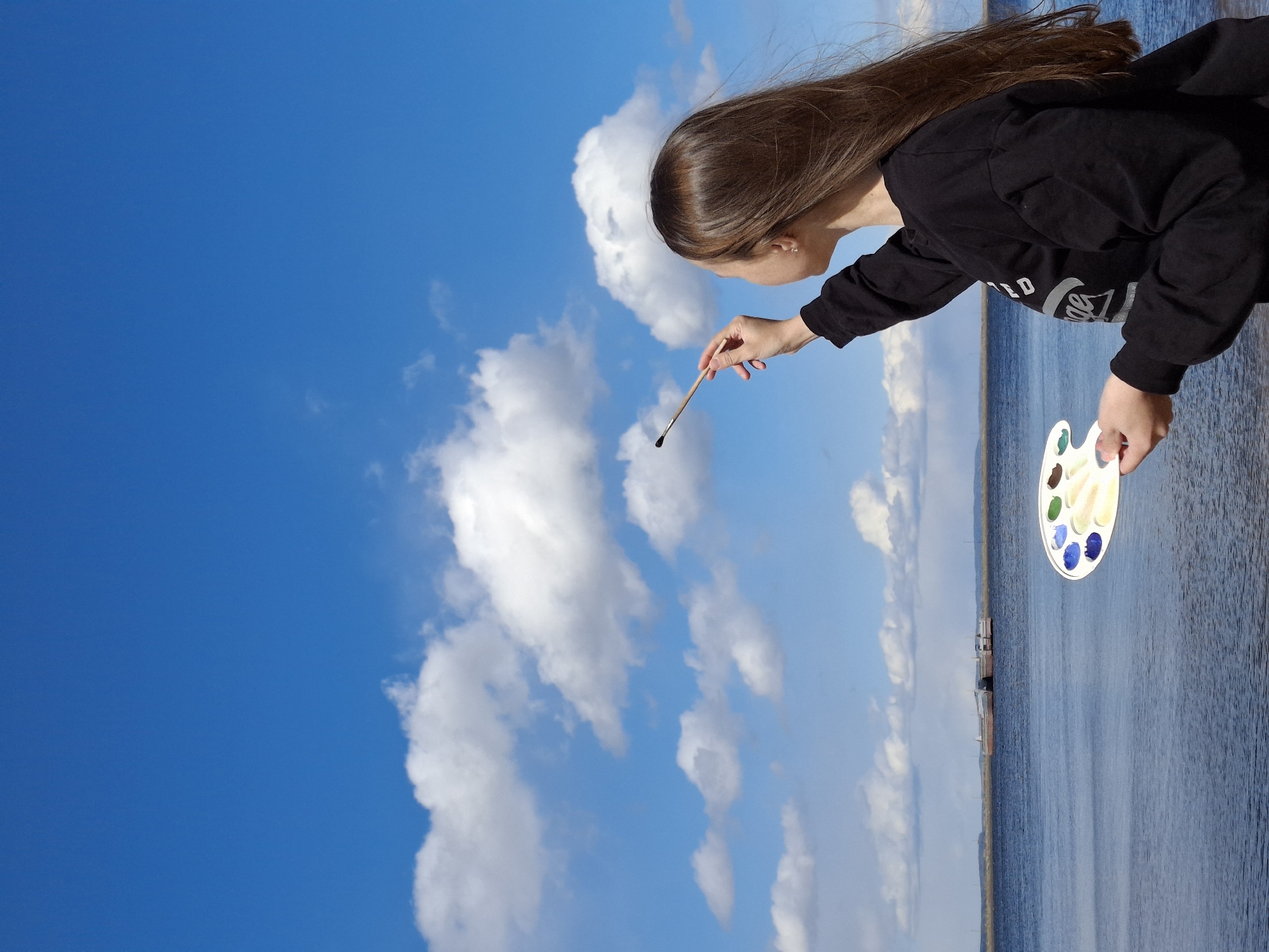 The image displays a young woman in an artistic pose, seemingly "painting" the sky, with the photo rotated 90 degrees counter-clockwise. The woman, with long brown hair, is wearing a black long-sleeved shirt with the "Coca-Cola" logo and other text visible. Her right hand holds a paintbrush, pointing upwards towards the expansive blue sky, while her left hand holds a white artist's palette with various blobs of paint, including blue, green, yellow, and white/brown.
The background features a vibrant blue sky filled with scattered white cumulus clouds. Below the sky, across the horizon, is a large body of calm water, likely a river or lake, stretching into the distance. A faint shoreline with what appear to be distant structures or boats is visible on the water's edge.
The activity depicted is a conceptual art act, where the woman interacts with the natural landscape as if painting it. The scene suggests a bright, sunny daytime, possibly with a gentle breeze implied by the slight movement of her hair. The setting is consistent with a riverside or lakeside location, such as the Kama River in Chaikovsky, Russia, as per the context.