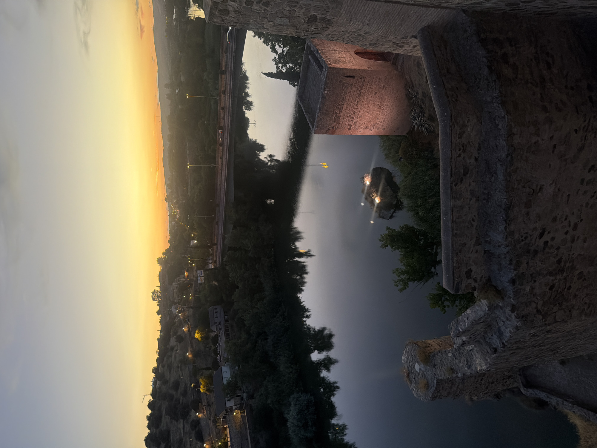 This is an elevated nighttime or twilight view of the Tagus River and surrounding landscape in Toledo, Spain. In the foreground, an ancient stone fortification, possibly part of the city walls or a castle, is visible on the right and in the lower left. The fortification features weathered stone blocks and a cylindrical turret-like structure on the right, which is illuminated by a warm, reddish light.
The Tagus River dominates the midground, its dark, still waters reflecting the subtle colors of the sky and the lights from the city. A low island or rock formation with lights on it sits in the river. A bridge, likely carrying vehicular traffic, spans the river in the distance, with cars visible crossing it. The banks of the river are lined with lush green trees and vegetation.
Beyond the river, the cityscape of Toledo unfolds. Buildings with illuminated windows are scattered across the hillside, and streetlights dot the landscape. The sky above displays a gradient of colors, transitioning from a pale yellow or orange near the horizon, where the sun has recently set, to a dusky blue or gray higher up. The overall atmosphere is serene and historic, capturing the timeless beauty of Toledo at dusk.