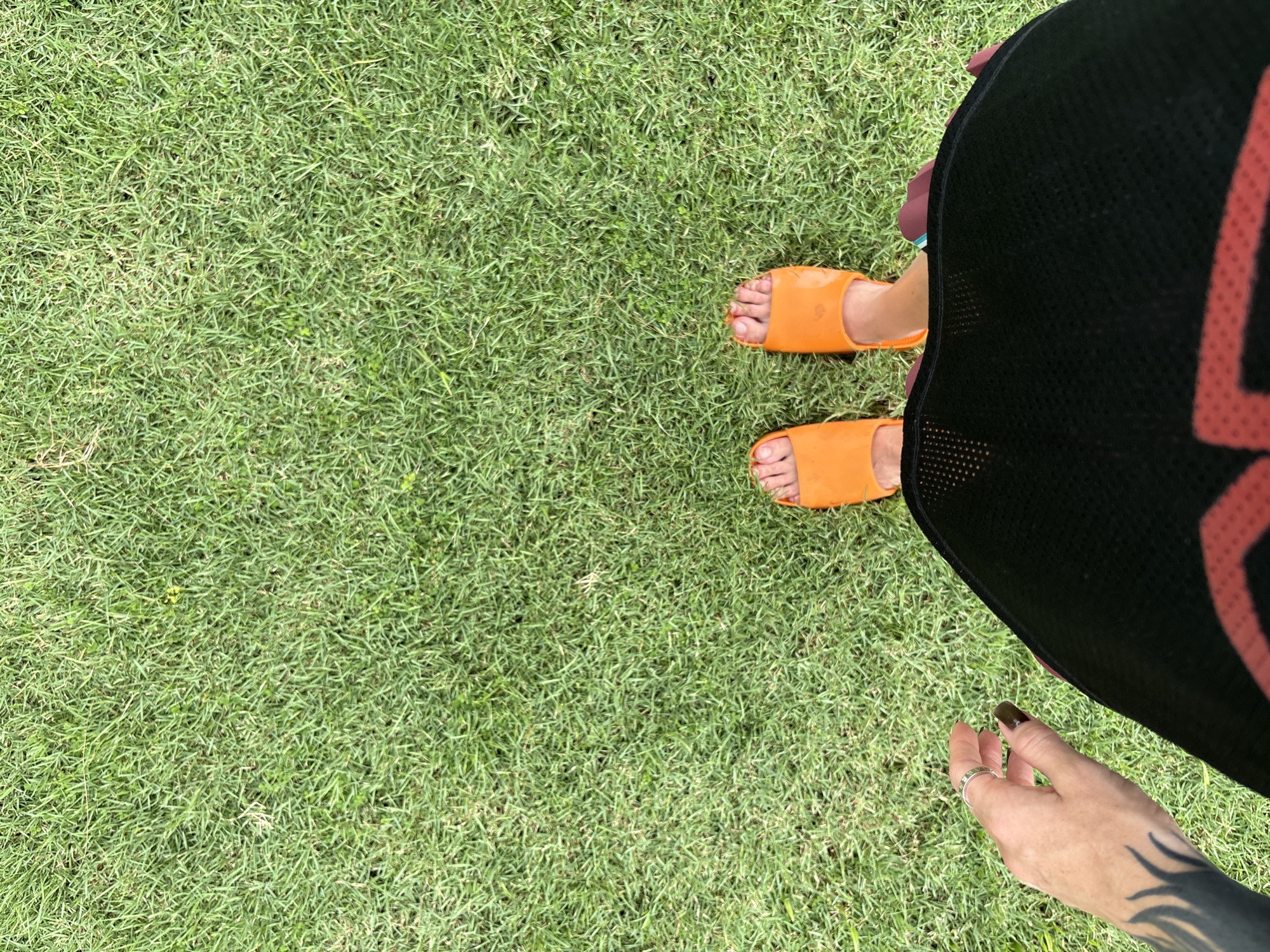 A person's feet, clad in bright orange slides, stand on lush green grass. The perspective is from above, looking down. The person is wearing a dark garment with a red design partially visible at the bottom of the frame. A hand with a ring and dark nail polish is visible on the left side of the image, resting near the garment. The scene appears to be outdoors, likely in Karon, Thailand, given the tropical-like grass.