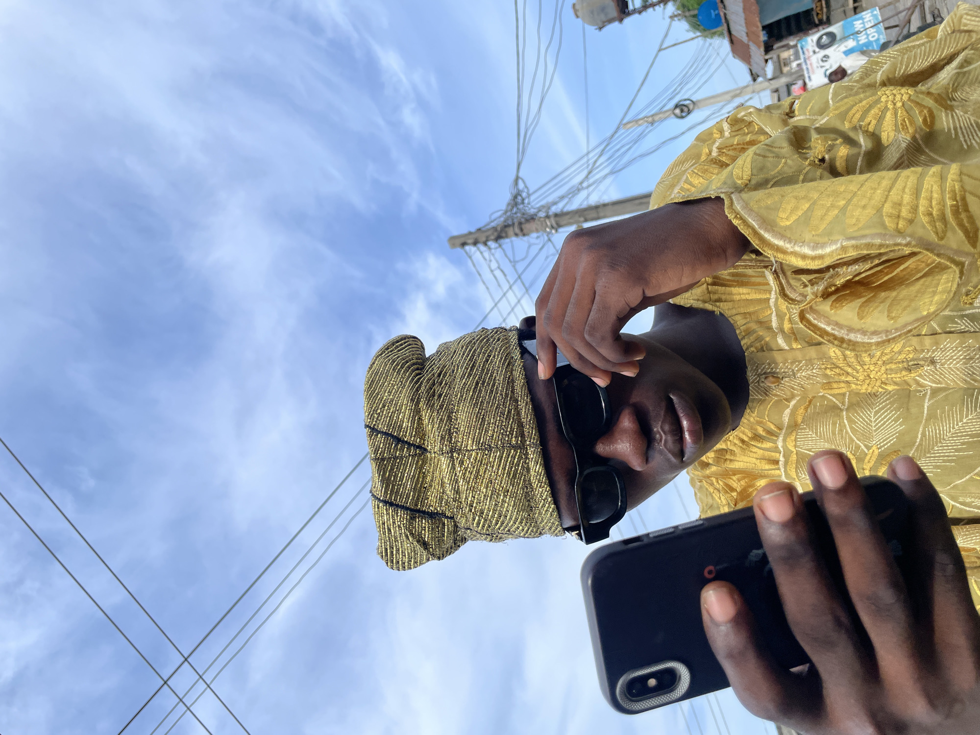 An image of a person wearing a golden-colored garment and headwrap with sunglasses, holding a black smartphone. The person appears to be standing outdoors in Ado, Nigeria, based on the presence of overhead power lines and a commercial building with a "NOW OPEN" sign in the background. The sky is partly cloudy.