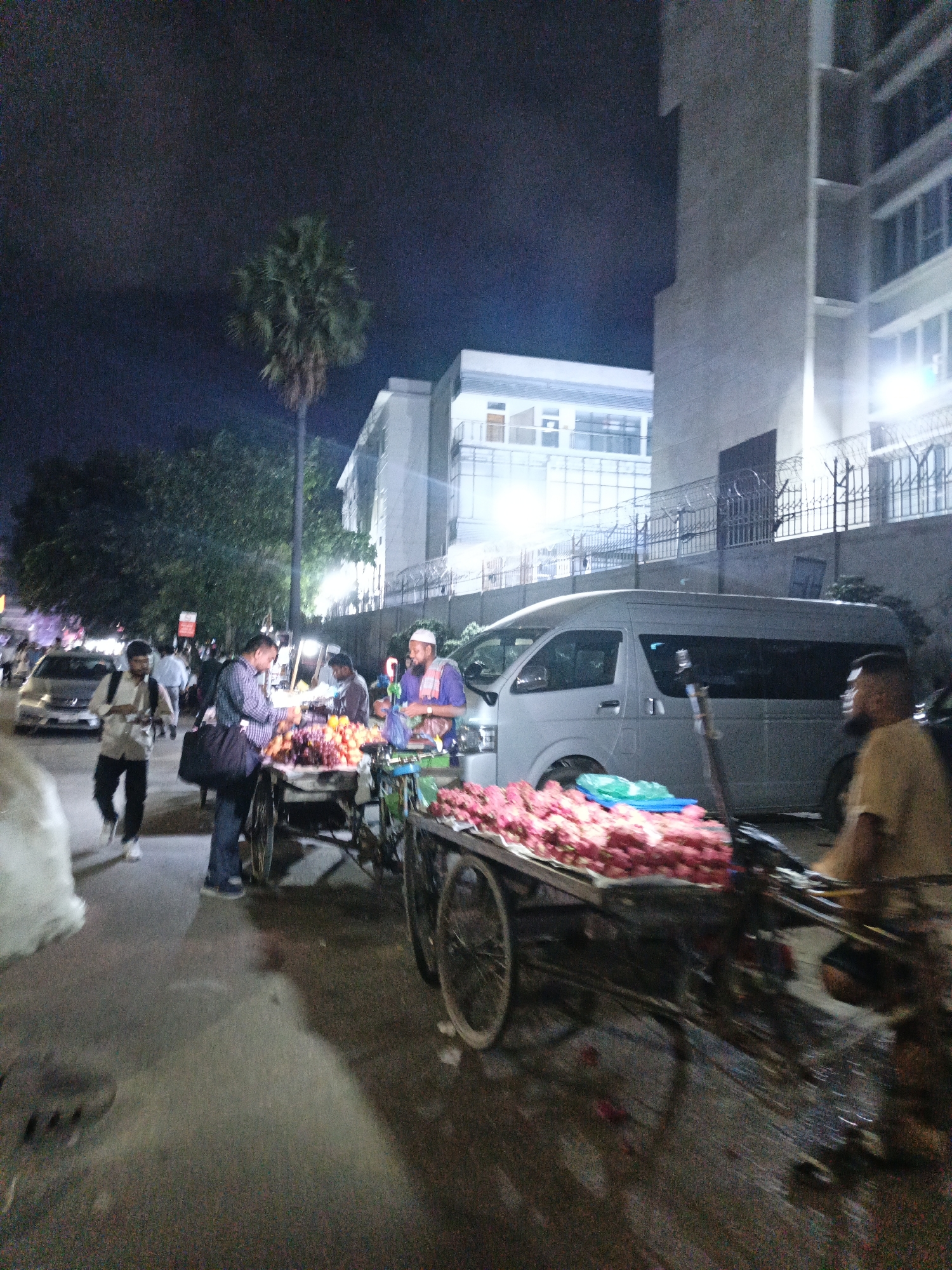 This is a night-time street scene in Dhaka, Bangladesh, likely a market area, featuring several individuals and street vendors. In the foreground, a fruit cart is laden with various fruits, including grapes and other produce, with a vendor wearing a purple tunic and a white cap standing behind it. To the left, another person is seen interacting with the fruit vendor, and further left, a man with a backpack walks on the street. A van is parked in the background, and a tall building with many windows is visible to the right. Trees and streetlights contribute to the ambient lighting of the scene. The ground appears wet, possibly from recent rain or street cleaning, with reflections of the lights.